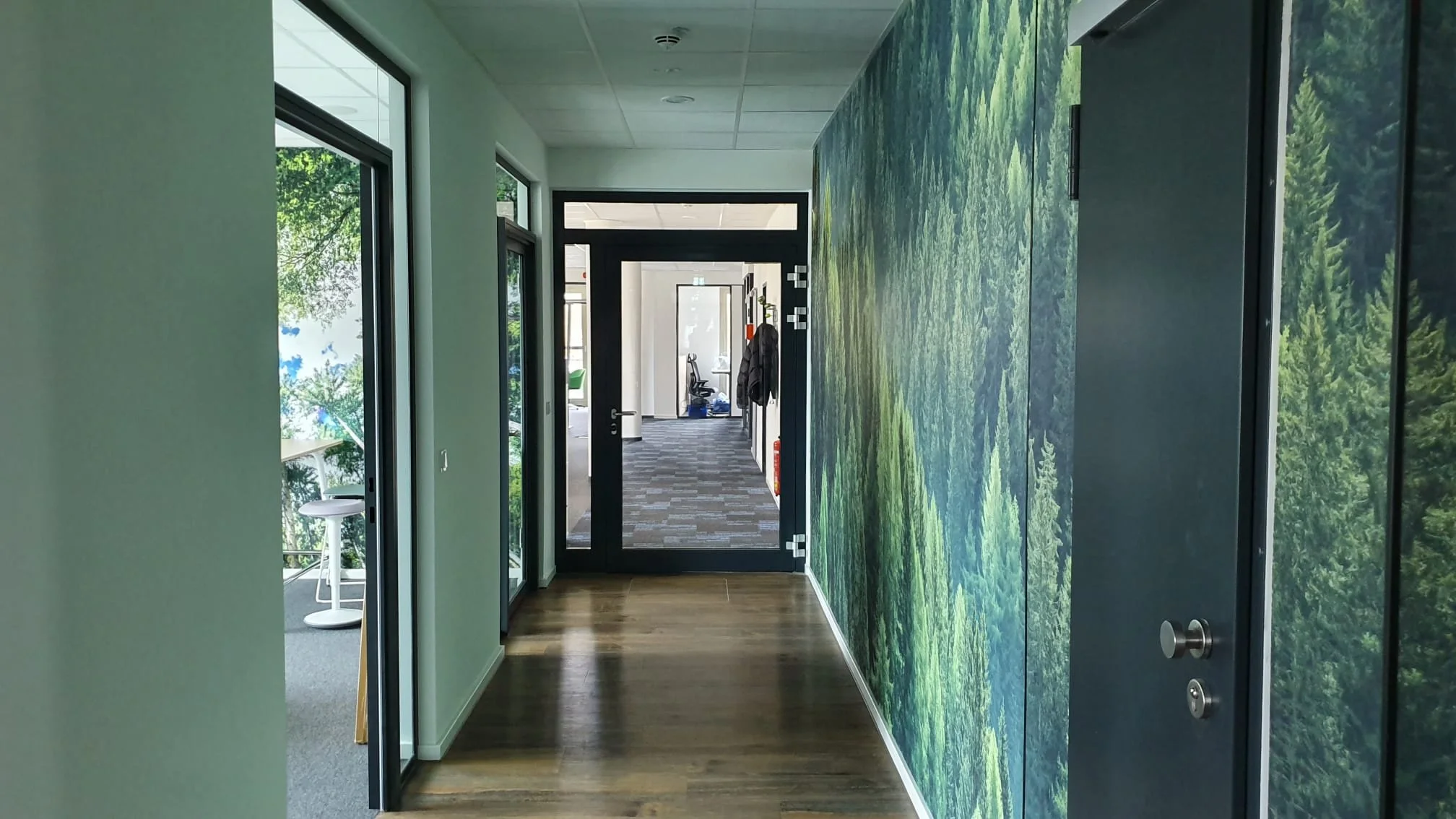 Innenansicht eines Flurs in einem modernen Bürogebäude mit Wanddekoration in Form eines grünen Waldes, Türen zu Bürozimmern auf beiden Seiten, und ein Raum am Ende mit Bürostühlen.
