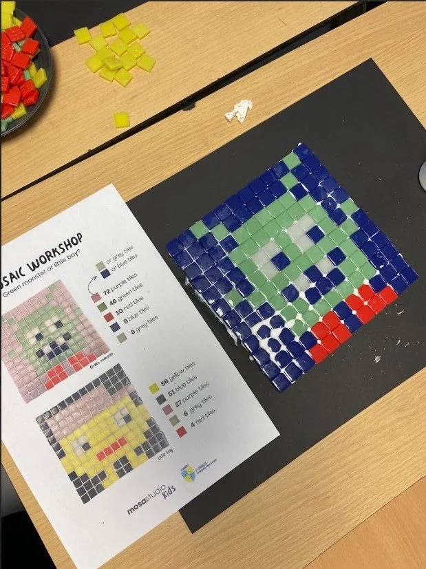 Mosaico en la mesa que representa un monstruo verde con ojos blancos y boca con dientes blancos, rodeado de fichas de colores y una hoja con instrucciones de mosaico.