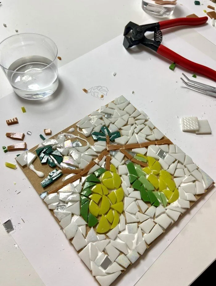Mosaico en proceso que representa un diseño de racimos de uvas con hojas y ramas, utilizando azulejos de cerámica en varios colores.