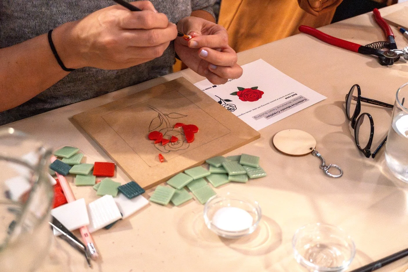 Persona creando un mosaico de una rosa utilizando piezas de cerámica en una mesa de trabajo, con herramientas y papeles alrededor. Team building original en Madrid. Taller de mosaico para empresas