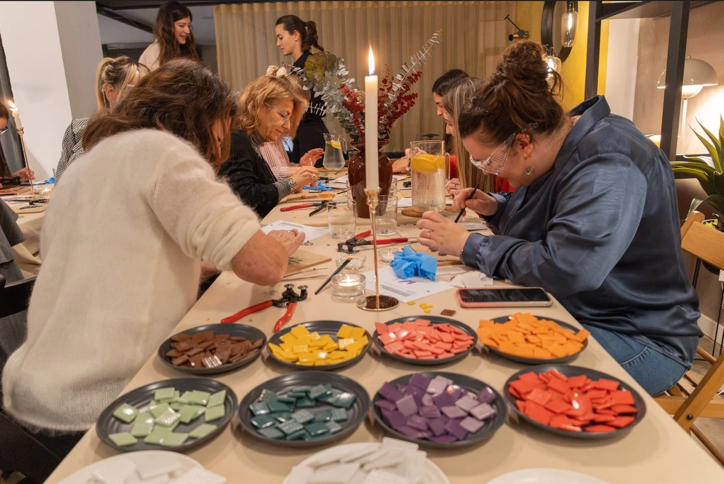 Grupo de personas participando en un taller de mosaico en una mesa decorada con múltiples piezas de azulejos de colores, herramientas, y líquidos en jarras con limón.