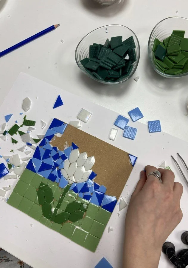 Persona armando un mosaico de flores blancas y verdes en una mesa, con tazas de mosaicos de diferentes colores y una pluma azul a un lado.