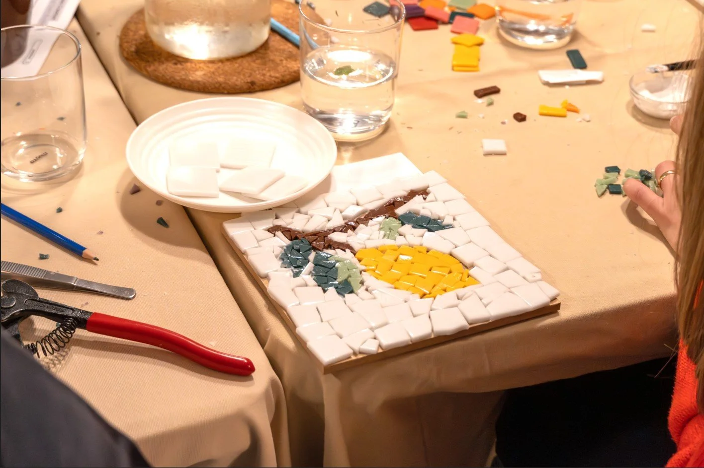 Mesa con mosaico en proceso, con piezas de azulejo de colores y herramientas de trabajo, como cortador y pinzas, en un entorno de taller creativo.