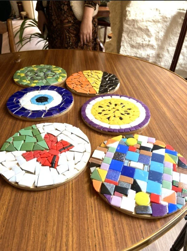 Seis platos decorativos con mosaicos de colores en una mesa de madera, con personas en el fondo.