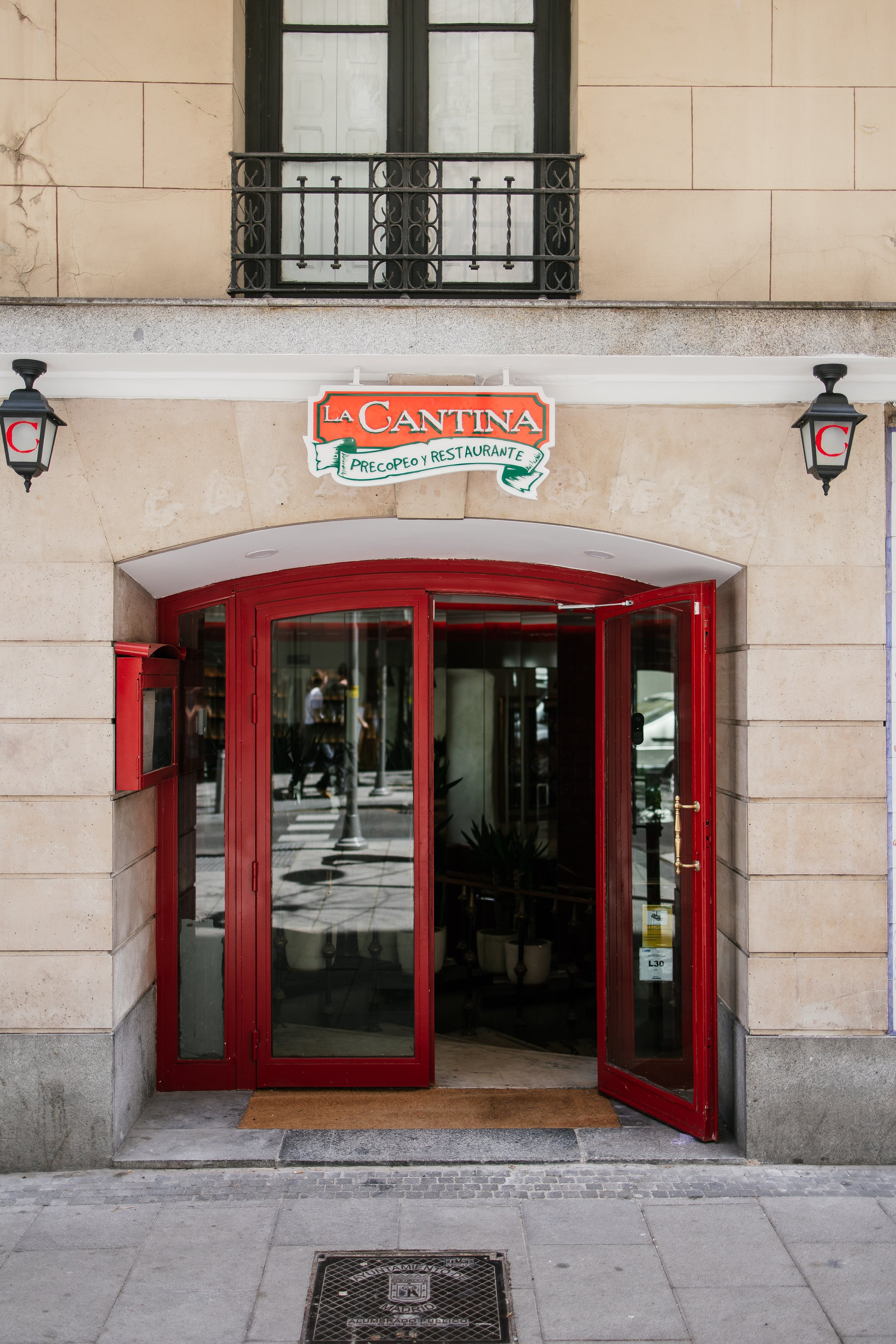 Entrada de un restaurante llamado La Cantina con puertas de color rojo, dos faroles negros en las paredes y un cartel que indica que es un restaurante y también un precopeo. La fachada es de piedra clara y tiene una ventana con barandilla negra y ventanas blancas.