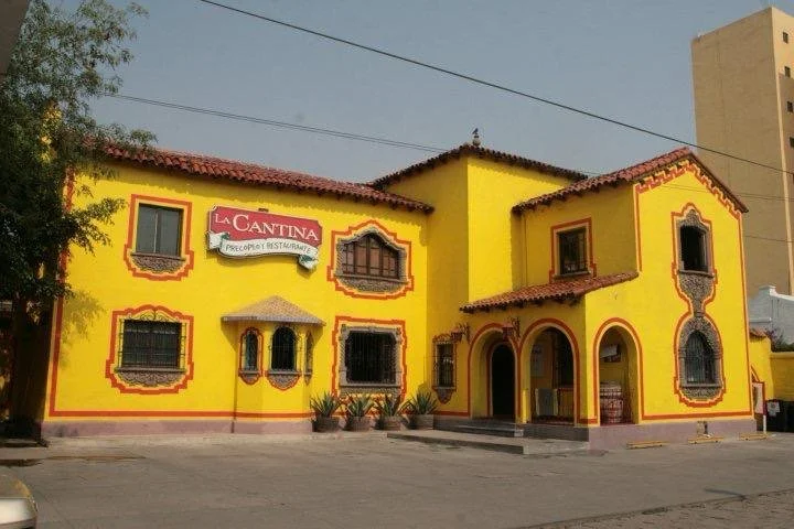 Edificio color amarillo con puertas y ventanas ornamentadas, cartel que dice "La Cantina" y áreas de entrada con arco, en una plaza.