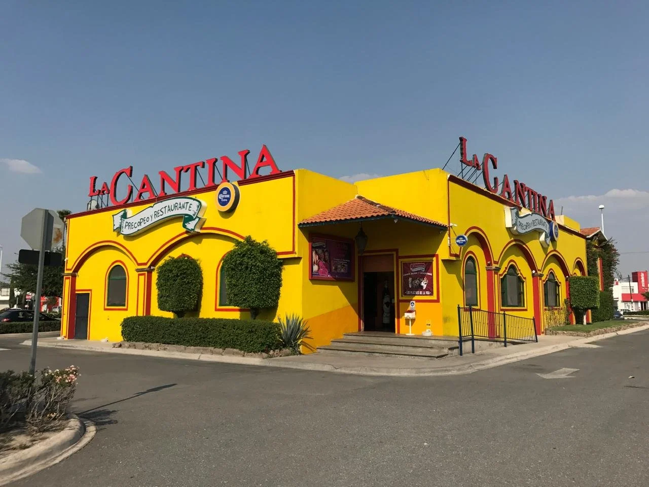Edificio de color amarillo con detalles rojos y señalización que dice 'La Cantina' y 'Precopeo y Restaurante', árboles decorativos y escalones en la entrada.