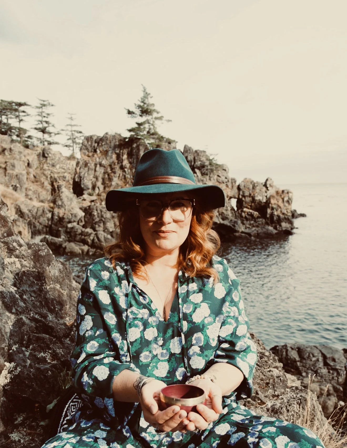 A woman with glasses and curly red hair sitting on rocky terrain near the water, holding a mug, wearing a wide-brimmed hat and a patterned dress with blue, green, and white colors.