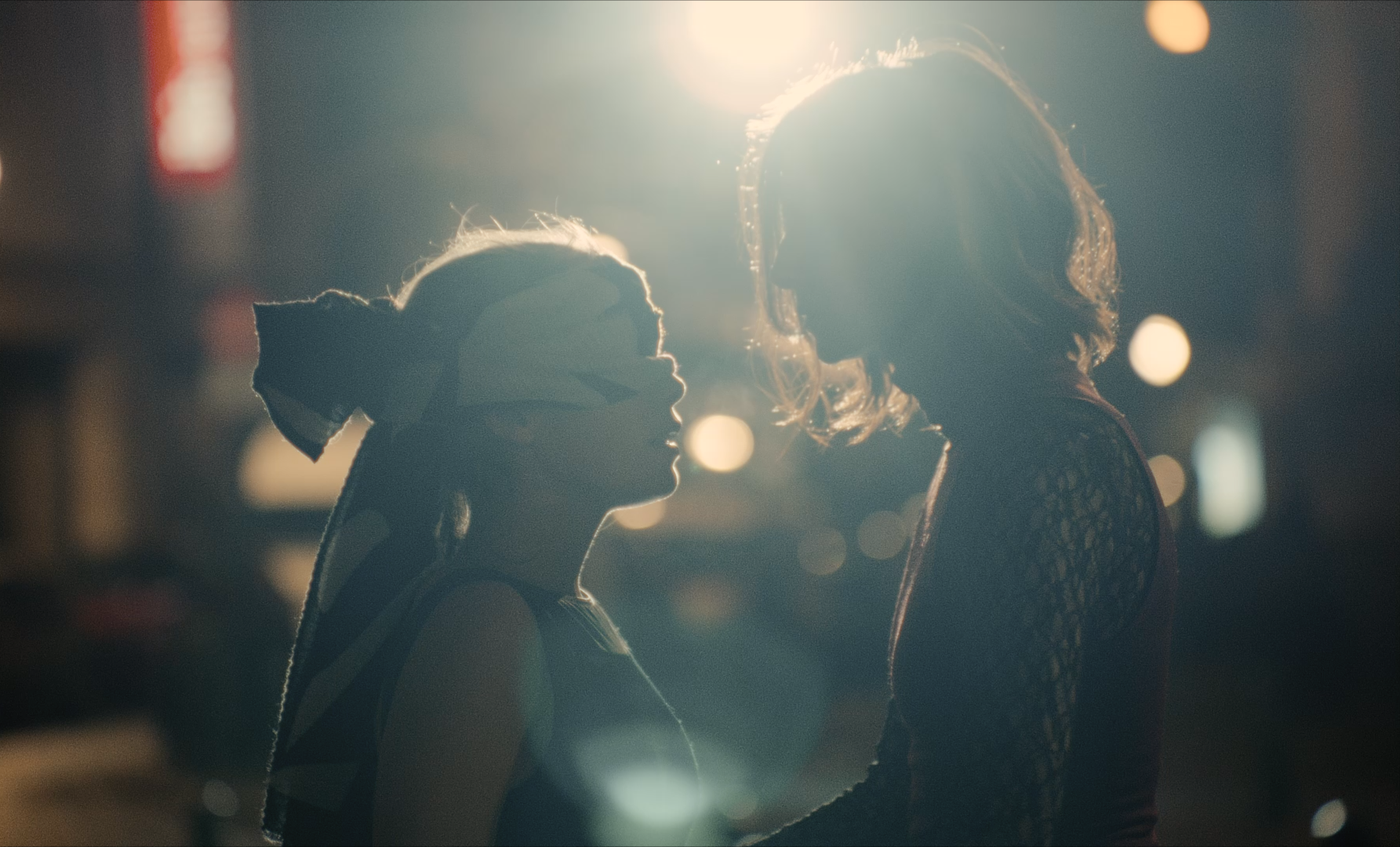 Two women face each other in a dimly lit setting with bright backlighting creating silhouettes and bokeh effects.