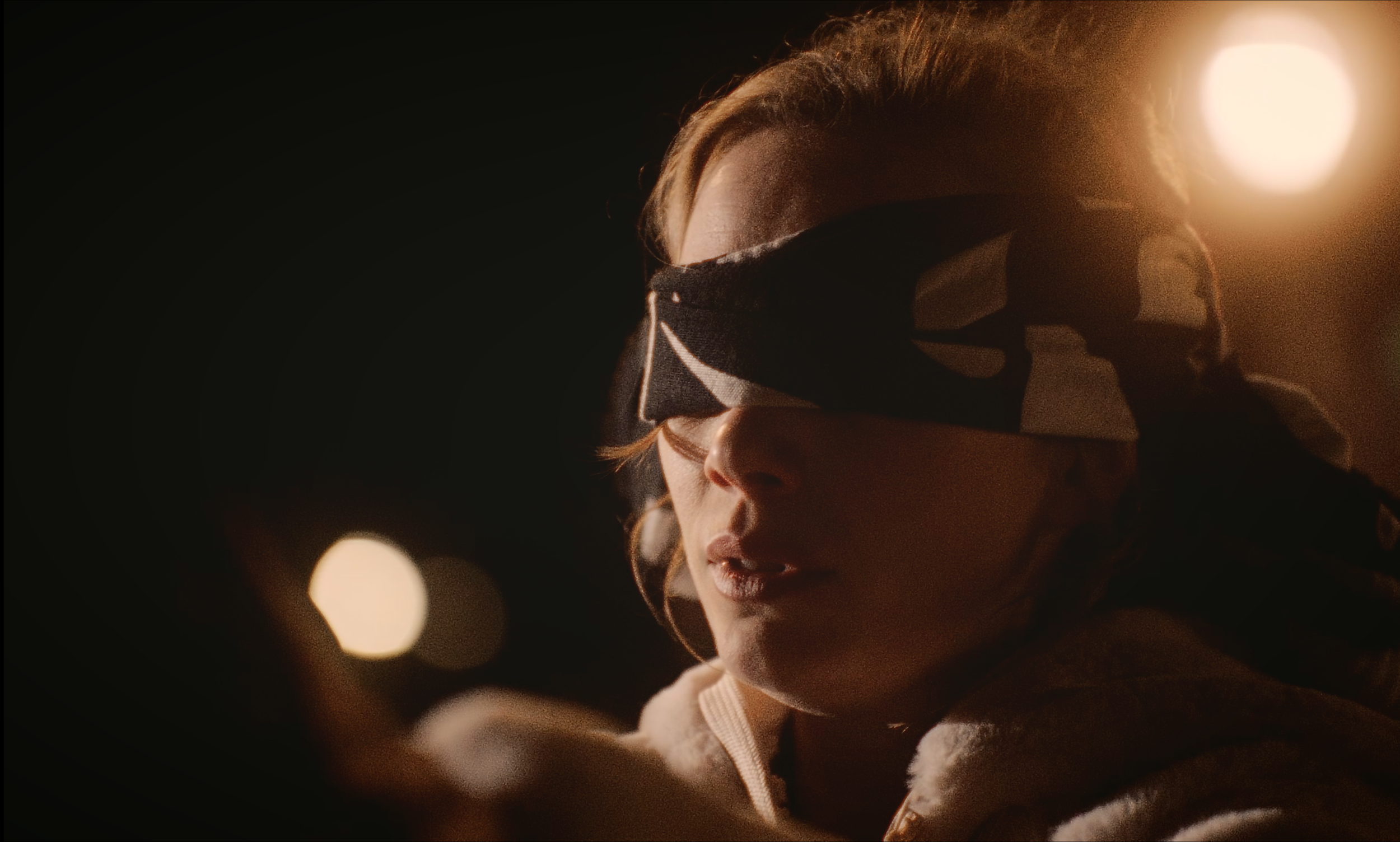 A woman with light brown hair and light skin wearing a black eye mask, illuminated by warm lighting, with blurred lights in the background.