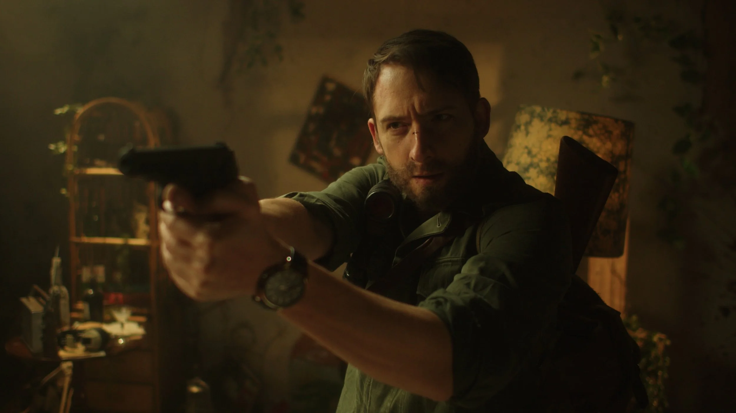 A man aiming a gun with a focused expression, wearing a watch and headphones around his neck, in a dimly lit room with shelves and plants in the background.