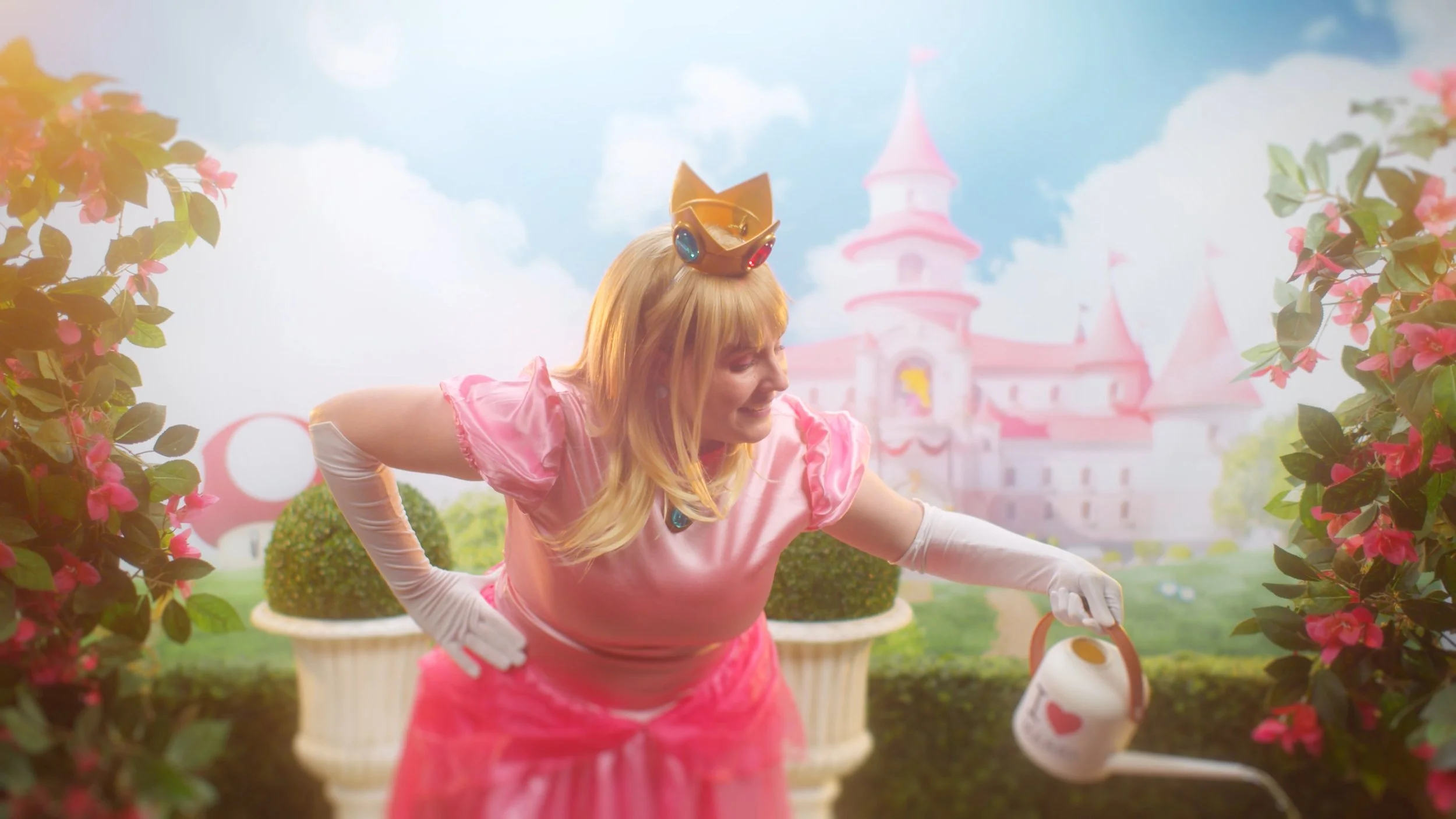 Person dressed as Princess Peach from Disney, smiling in front of a castle backdrop, surrounded by pink flowers, watering plants.