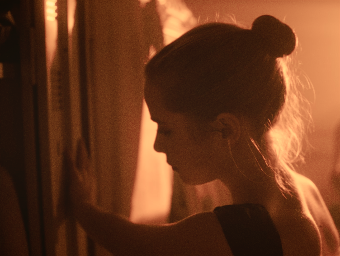 Silhouette of a woman with a bun hairstyle, looking down, in warm, orange lighting, standing near a wall.