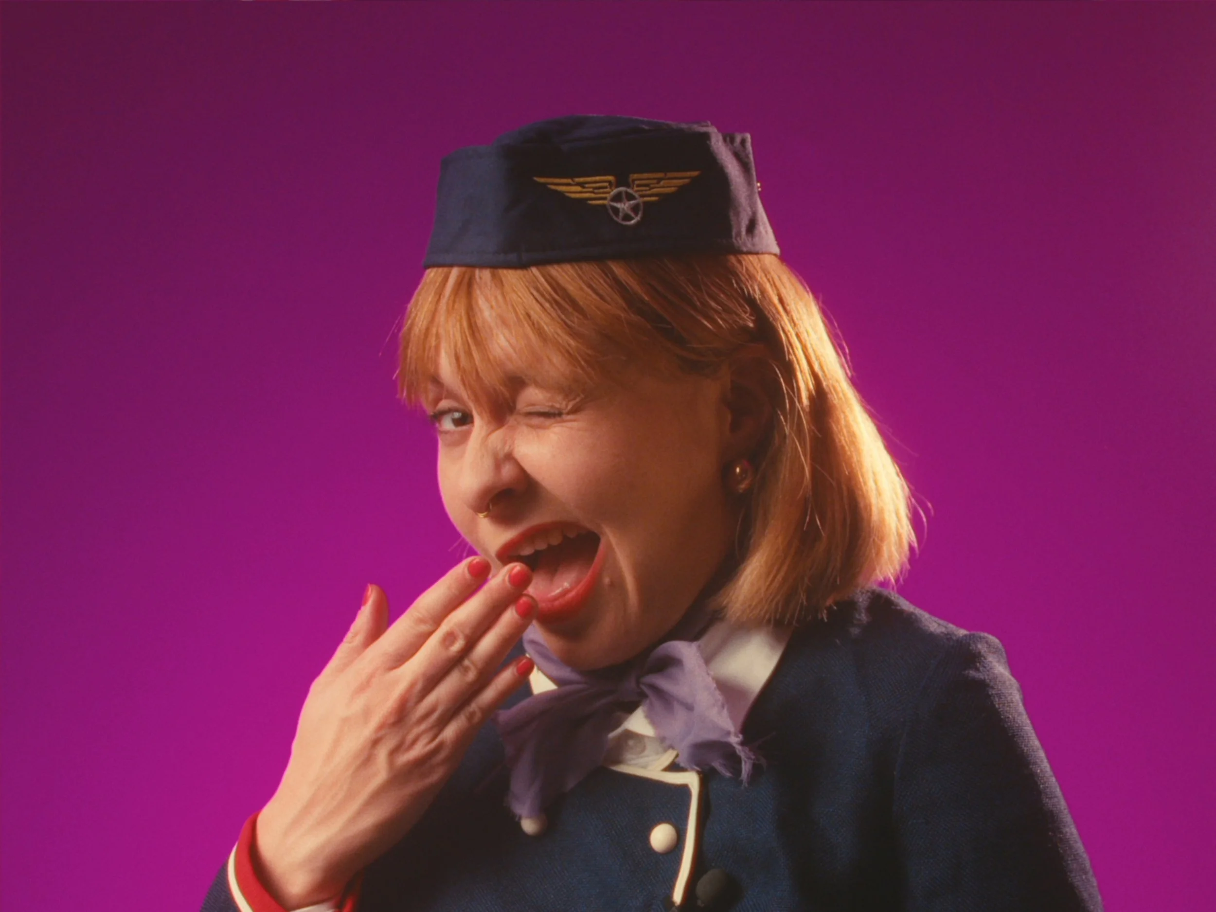 A woman with orange hair, wearing a navy cap with an aviation badge, winking with her mouth open and her hand near her face, against a purple background.