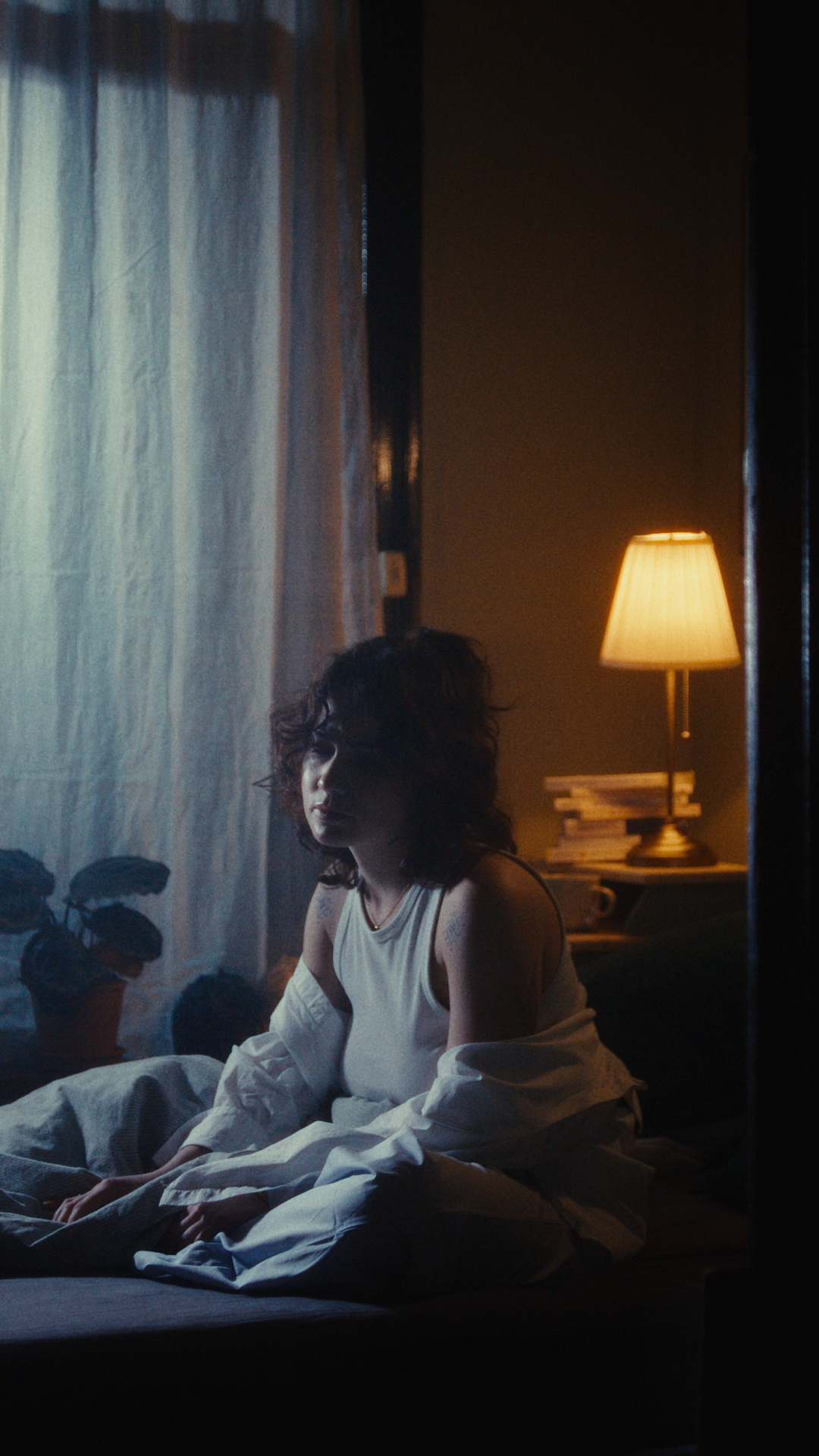 A woman with short dark curly hair sits on a bed in a dimly lit room. She is wearing a sleeveless white top and is looking slightly to the side. The room has a window with sheer curtains, a bedside table with a stack of books, and a lamp emitting war