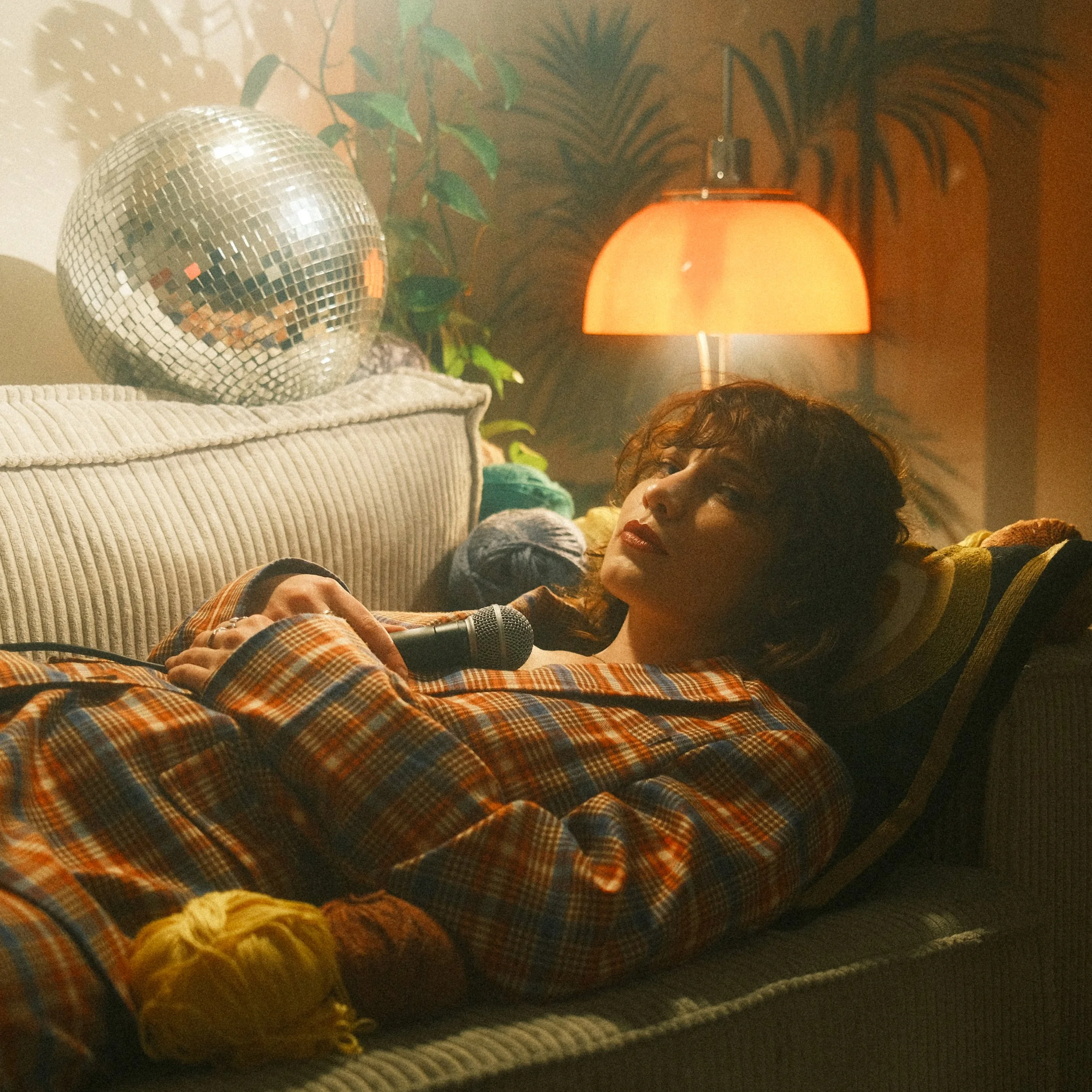 Close-up of a wafting table or shelf with a disco ball, green plants, a warm orange lampshade, a person with curly hair, and some yellow flowers.