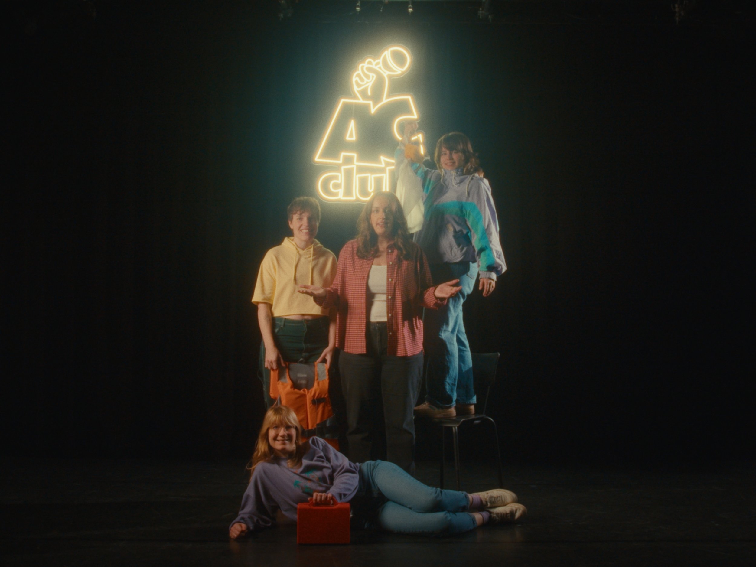 Group of five children on stage, one child standing and holding a glowing light, the others seated or lying down, with a neon sign of a hand holding a microphone and the words 'A club' in the background.