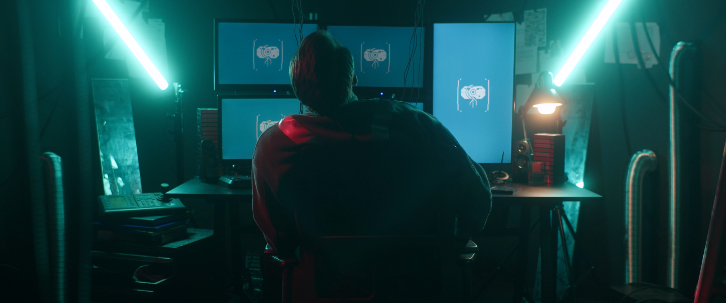 Person working in a dimly lit room with multiple computer monitors displaying a robot icon, illuminated by blue-green neon lights.