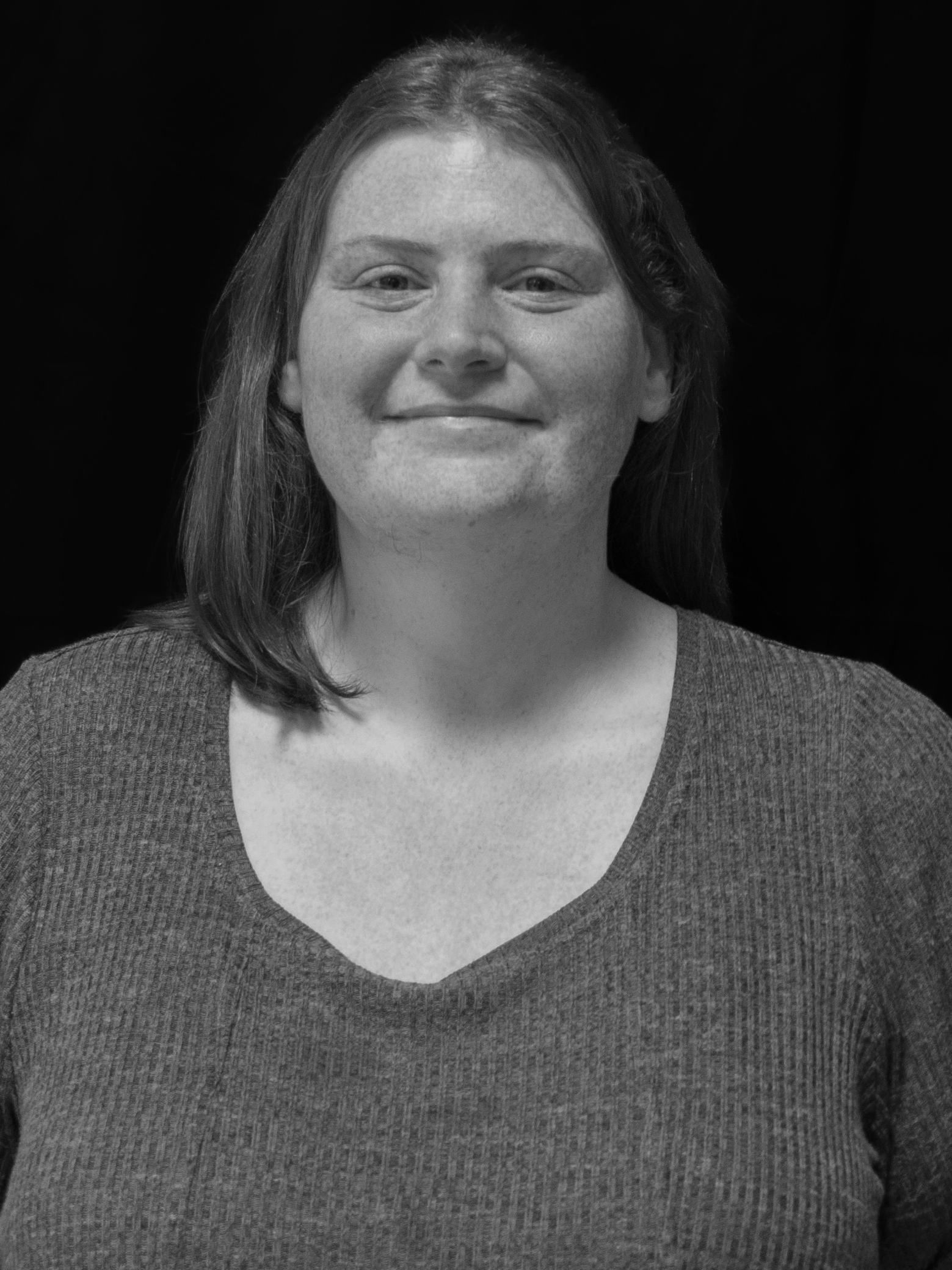 Black and white portrait of a smiling woman with shoulder-length hair, wearing a V-neck top, against a dark background.