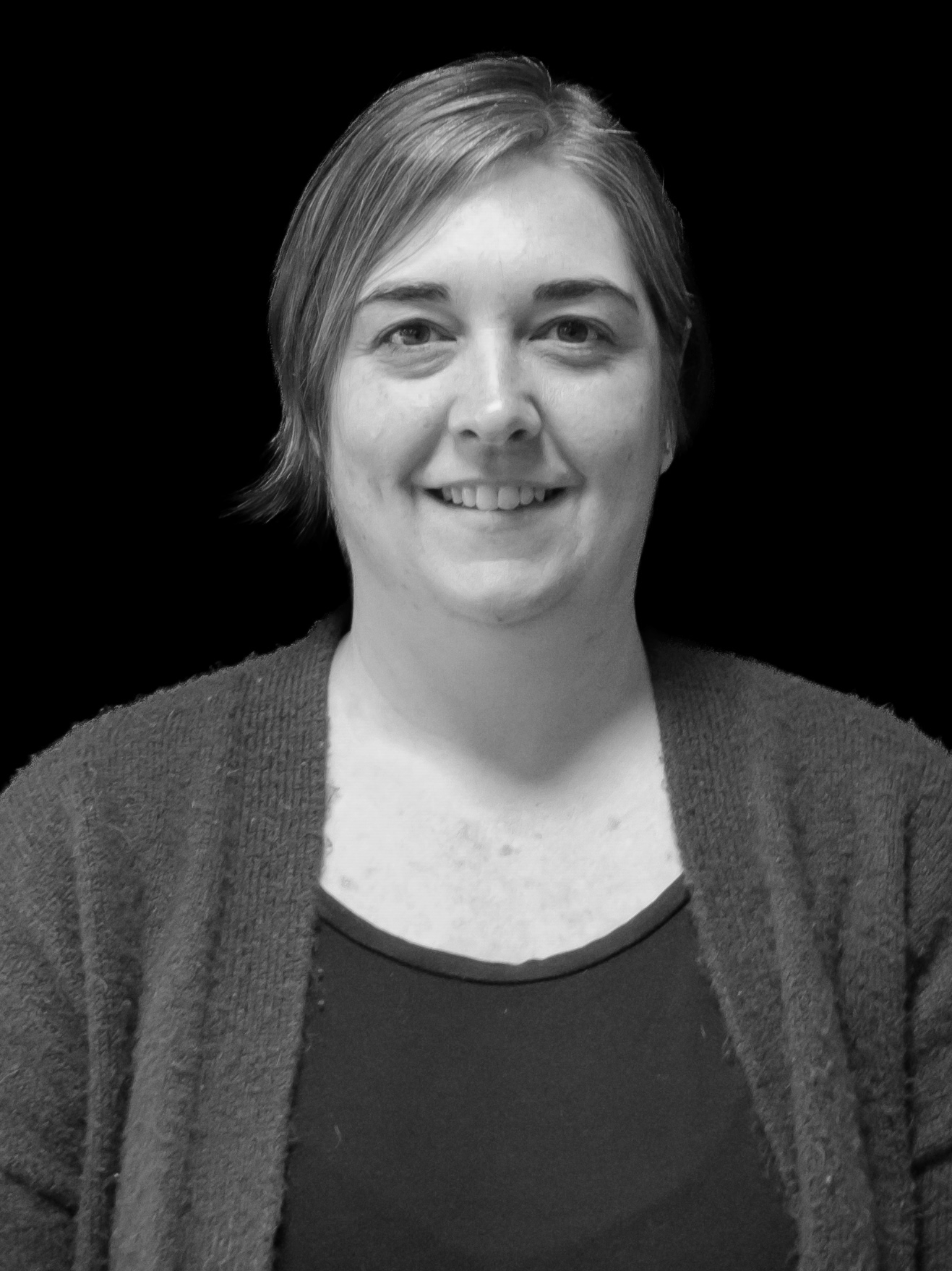 Black and white photo of a woman with short hair, smiling, wearing a dark top and a cardigan, against a black background.