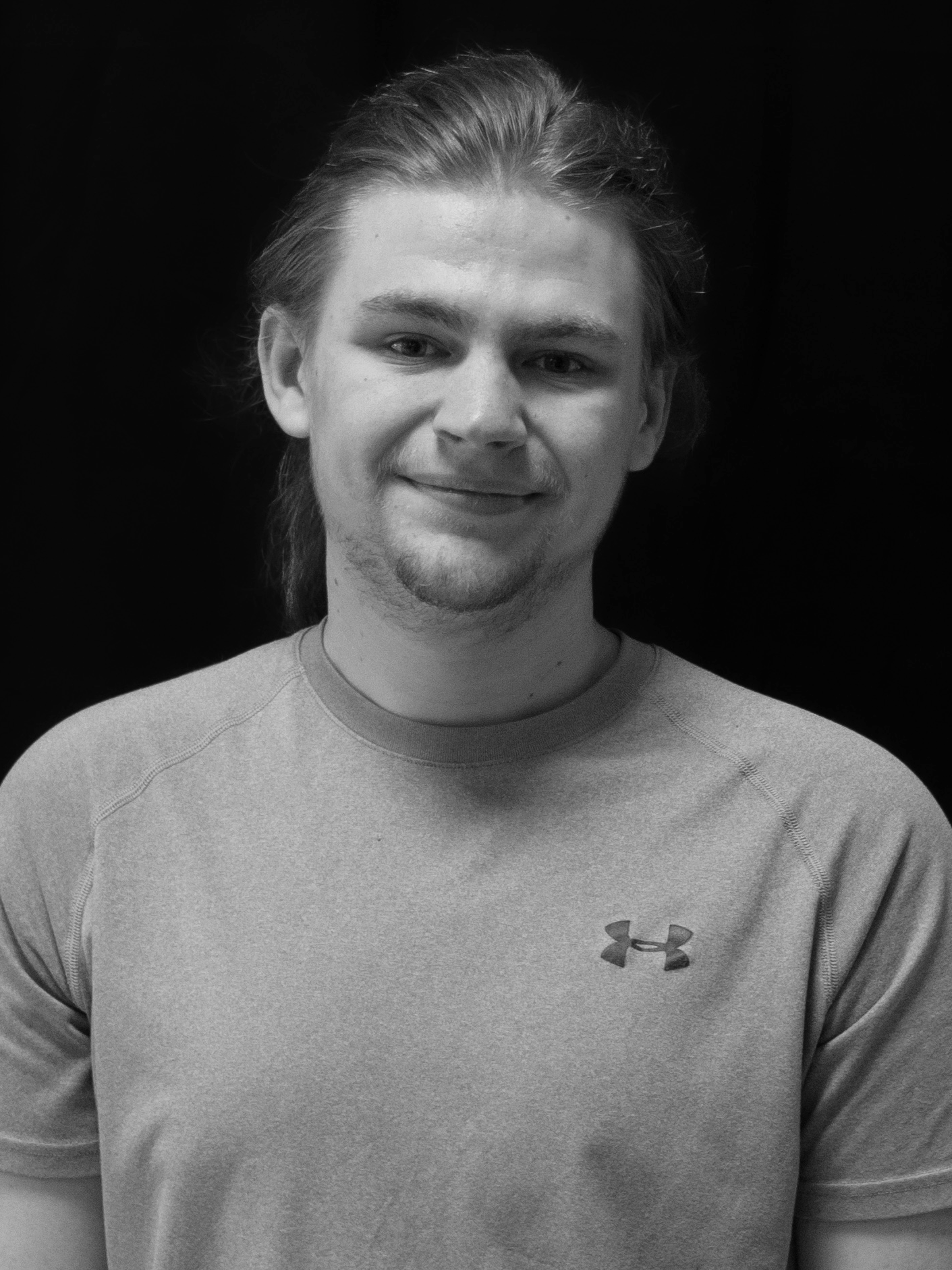 Black and white portrait of a young man with shoulder-length hair, smiling, wearing a sports shirt with an Under Armour logo.
