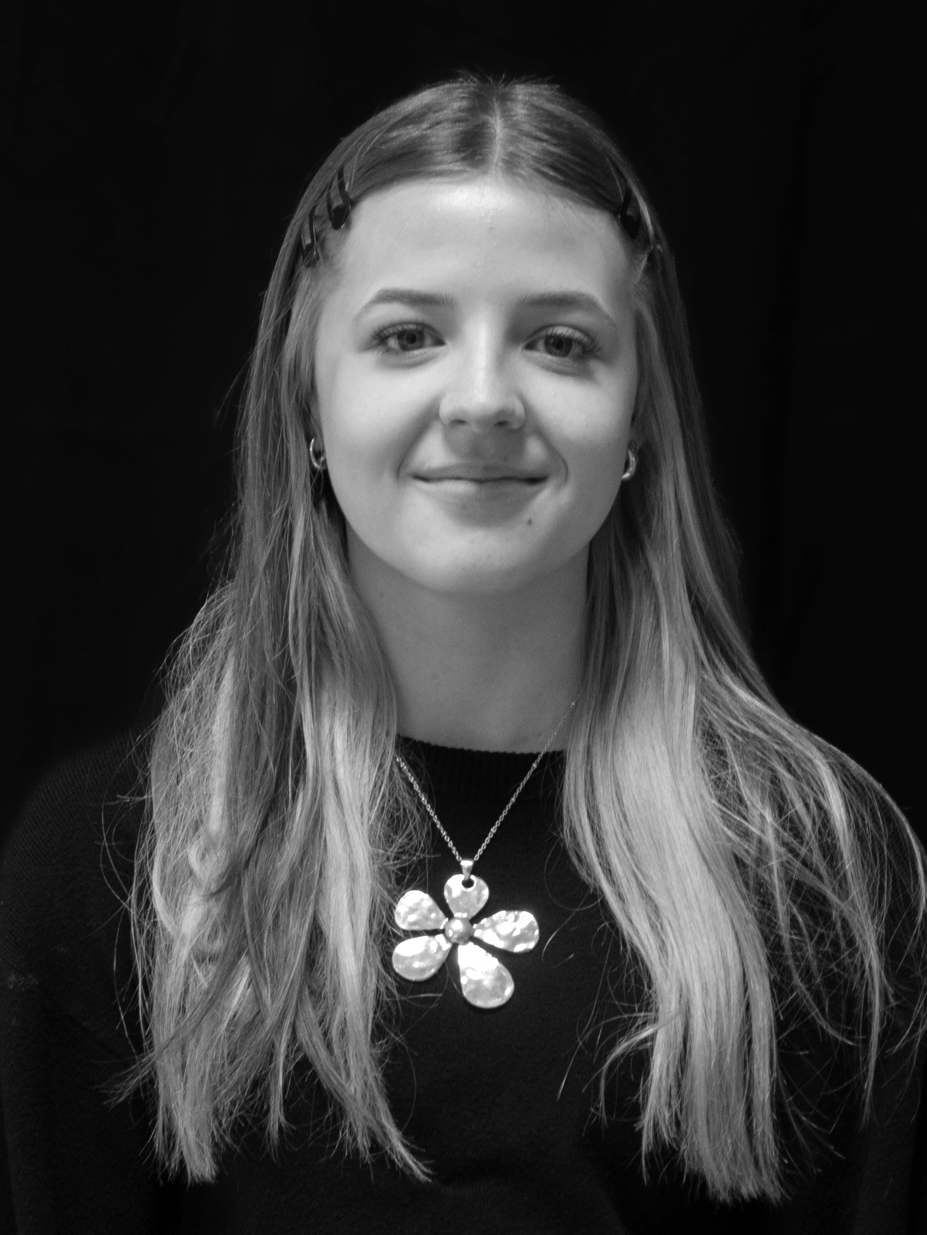 Black and white portrait of a young woman with long hair, wearing a necklace with a flower-shaped pendant and earrings, smiling softly, against a dark background.