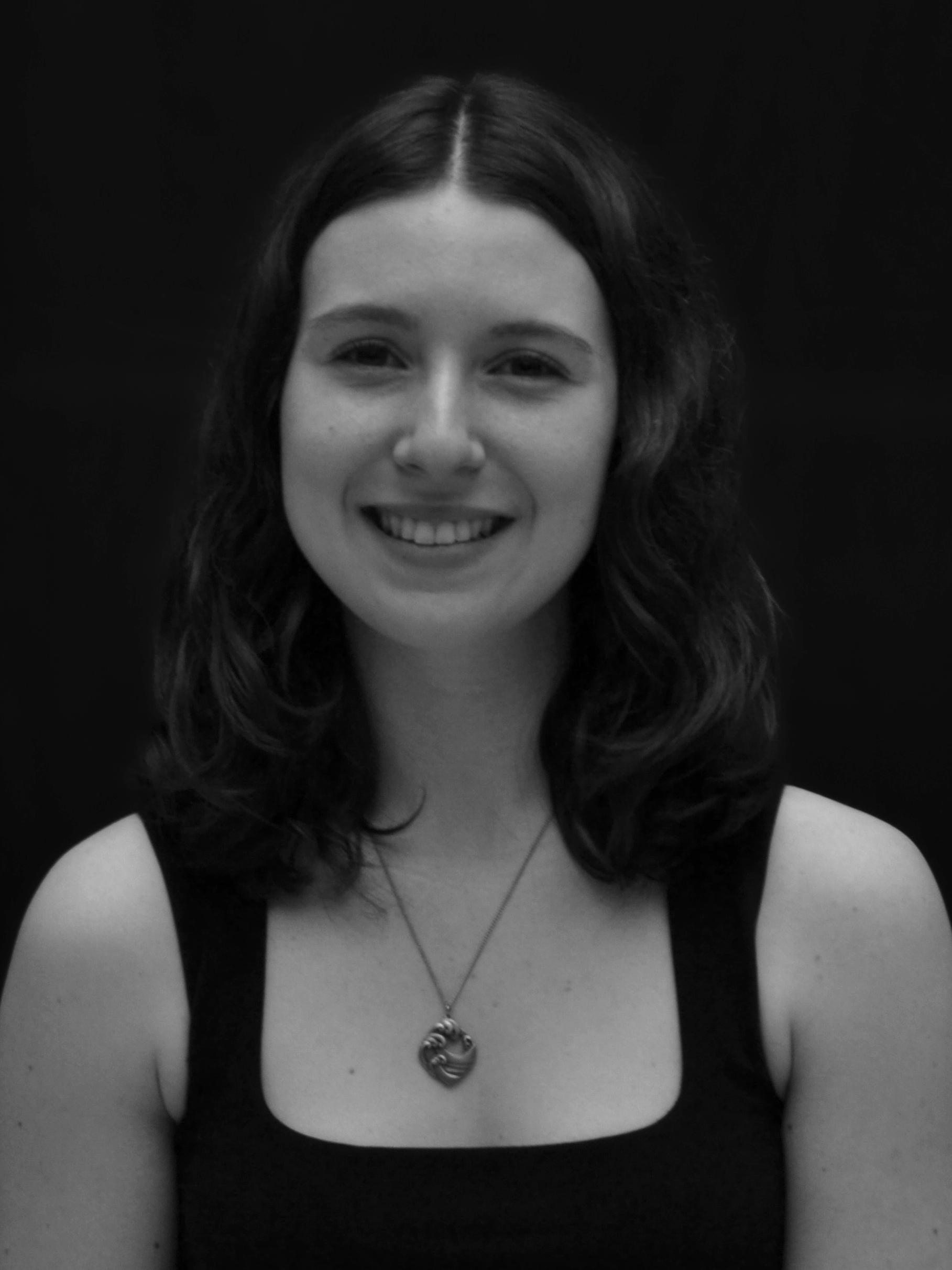 Black-and-white portrait of a young woman with shoulder-length wavy hair, wearing a sleeveless top and a necklace with a heart-shaped pendant, smiling at the camera against a dark background.