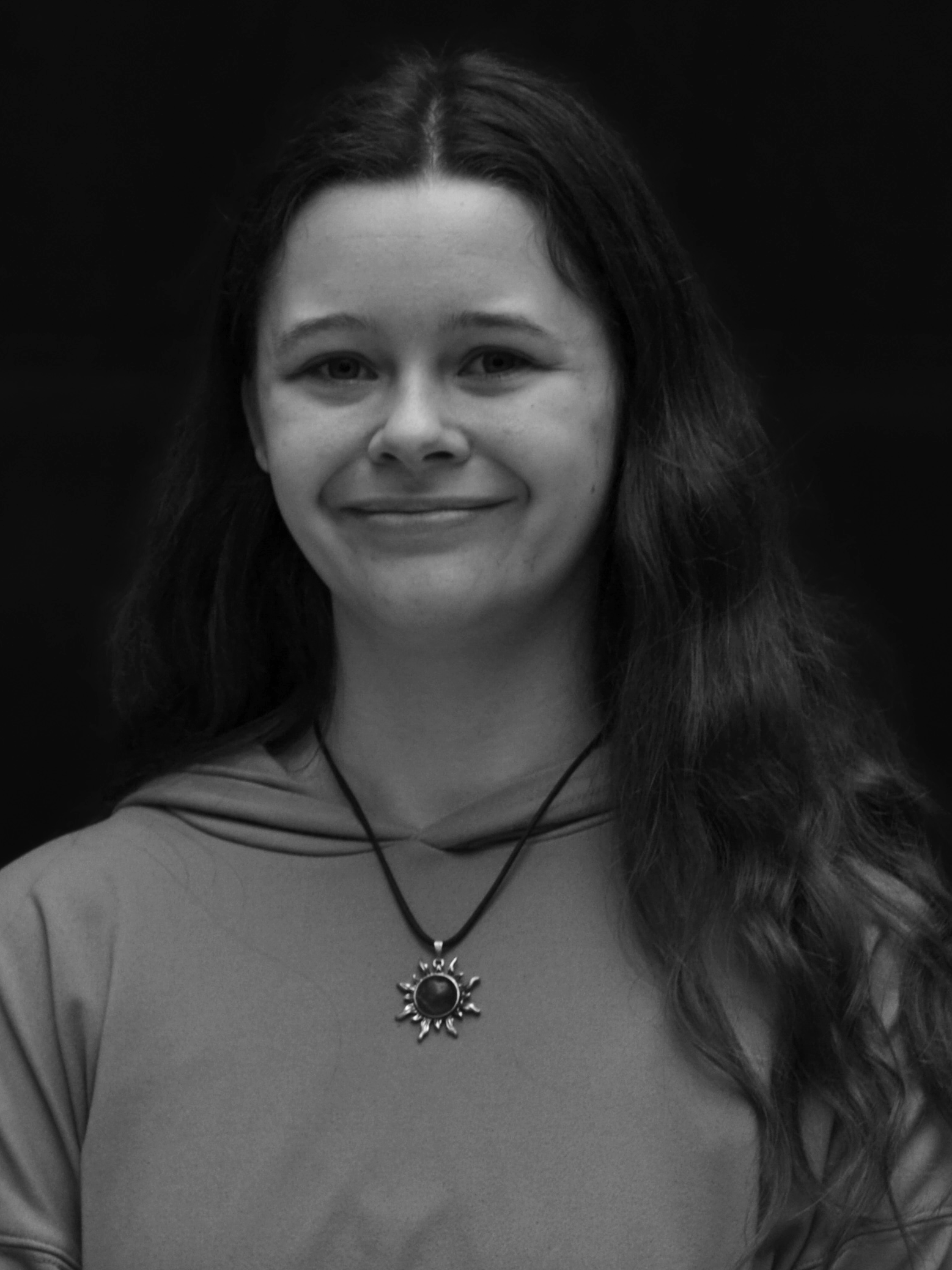 Black and white portrait of a young woman with long, wavy hair, smiling, wearing a necklace with a sun-shaped pendant and a hooded top.