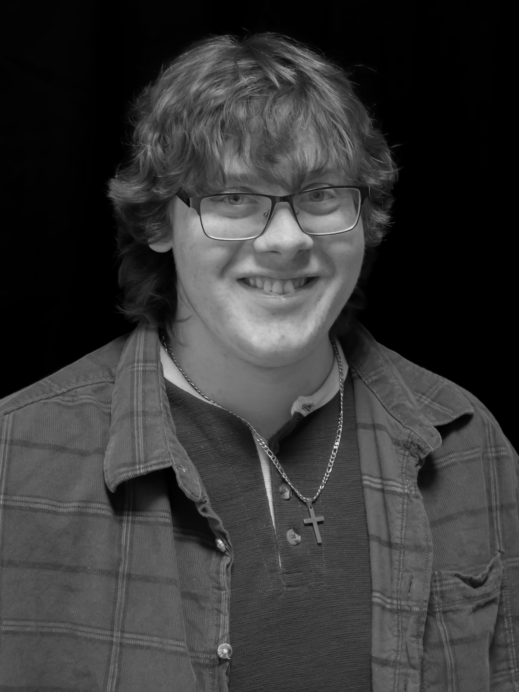 Black and white photo of a young man with wavy hair, glasses, and a friendly smile, wearing a collared shirt, a chain with a cross pendant, and a jacket, against a dark background.