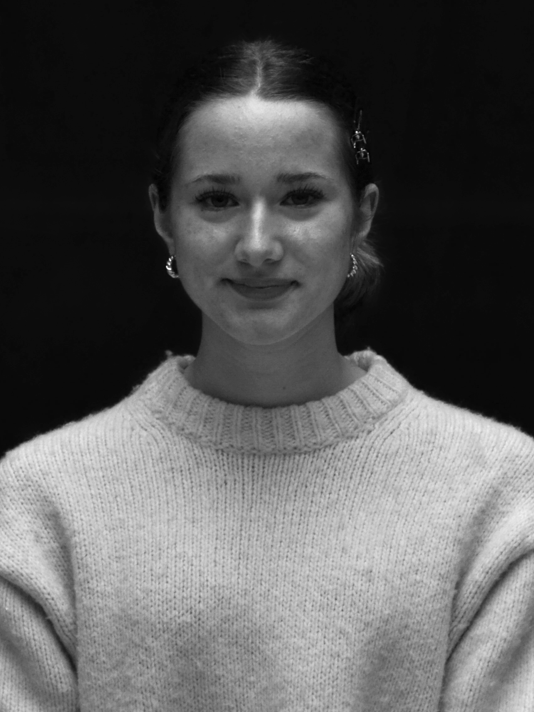 A woman with dark hair and earrings, wearing a light-colored knitted sweater, smiling slightly against a dark background.