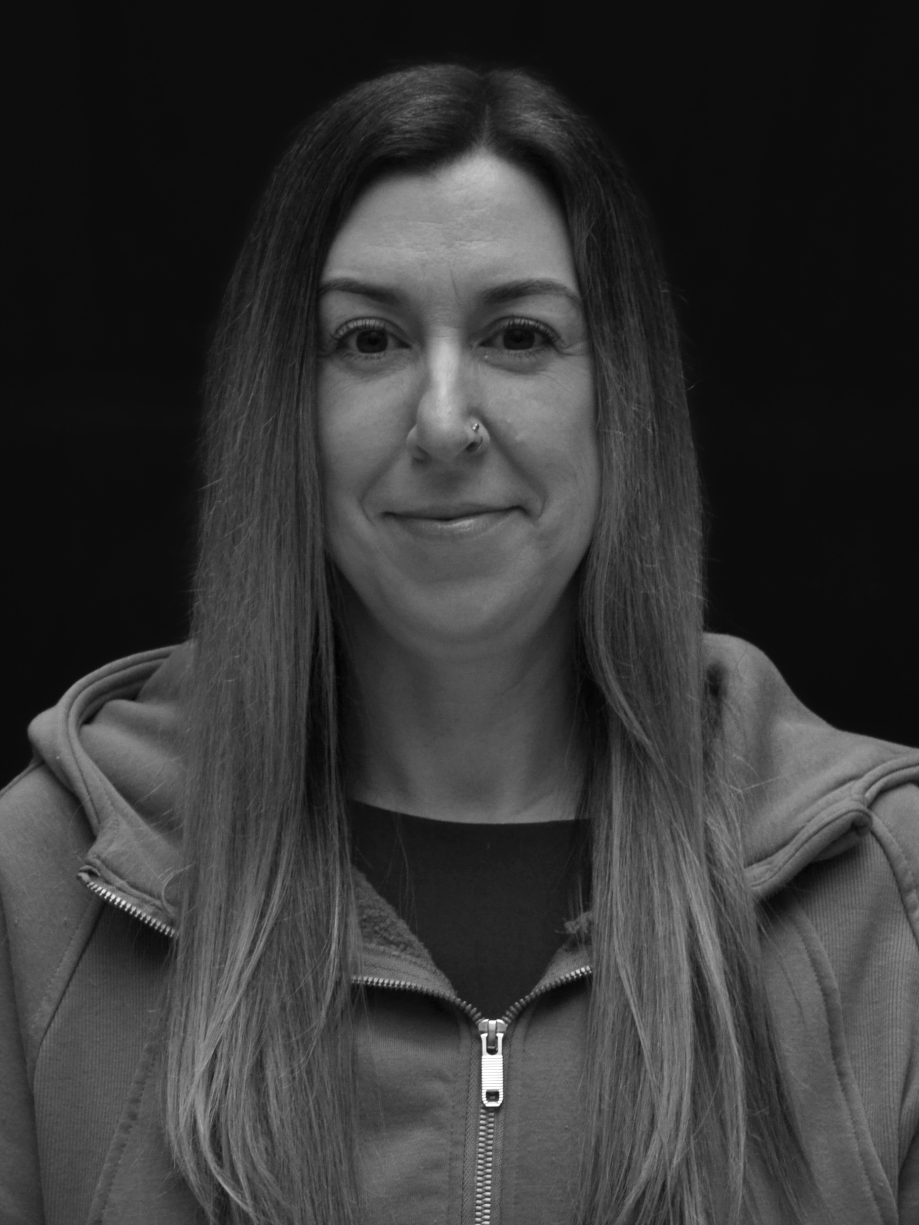 A woman with long hair, smiling slightly, wearing a zip-up hoodie and a nose piercing, against a dark background.