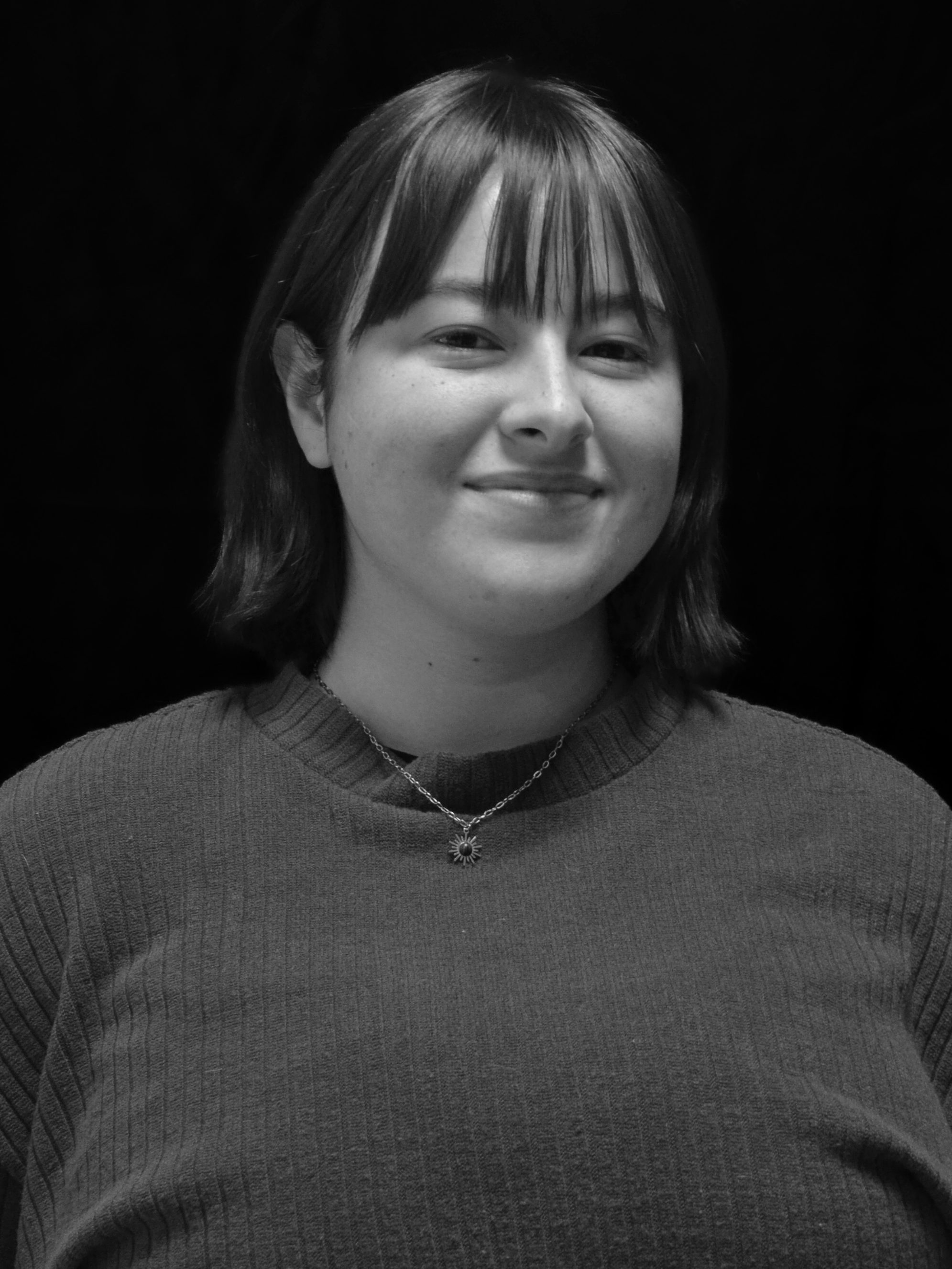 A young woman with shoulder-length hair smiling, wearing a sweater and a necklace with a sun-shaped charm, against a black background.