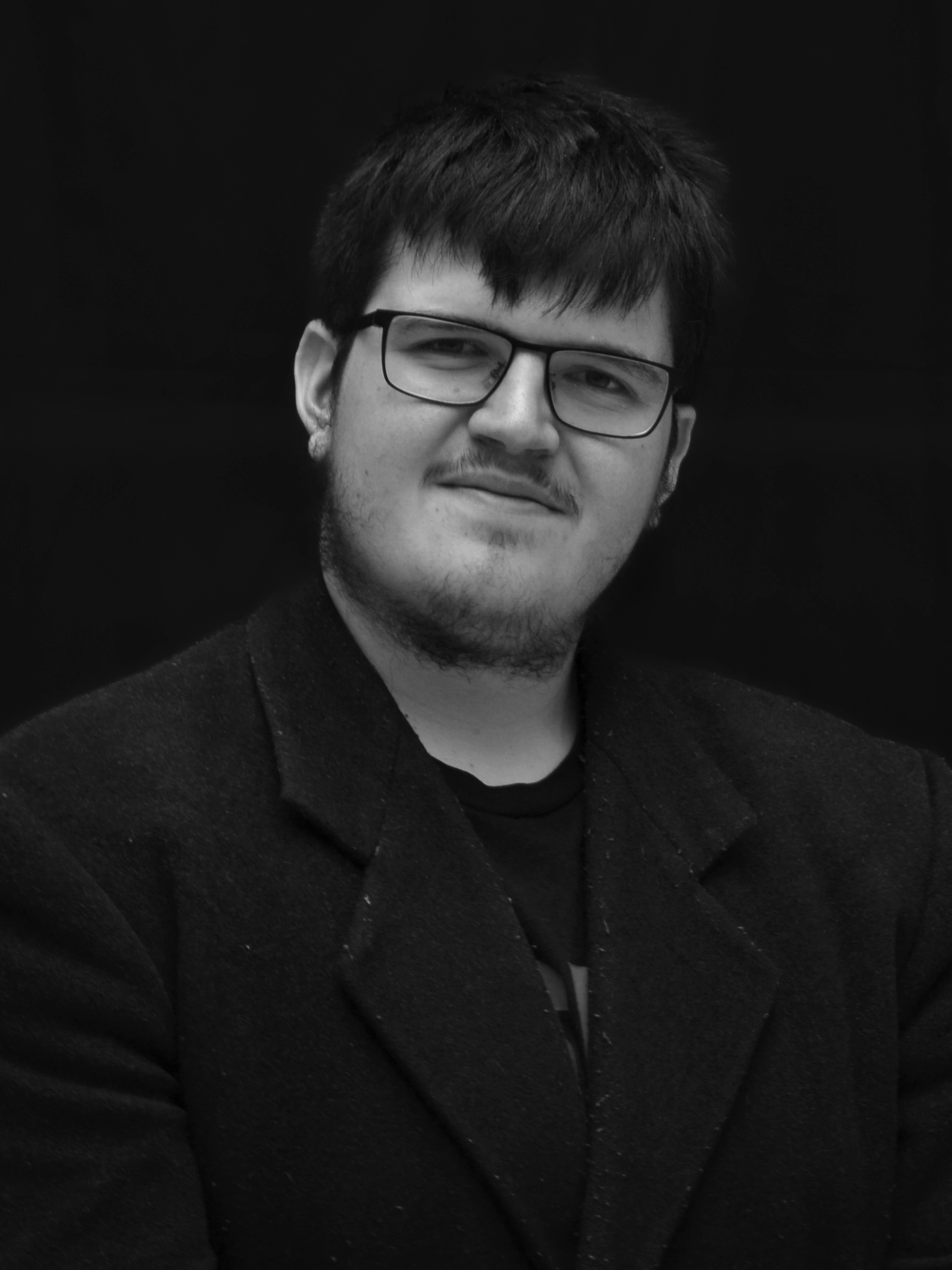 Black and white portrait of a young man with glasses, slightly smiling, wearing a dark blazer and black shirt, against a dark background.