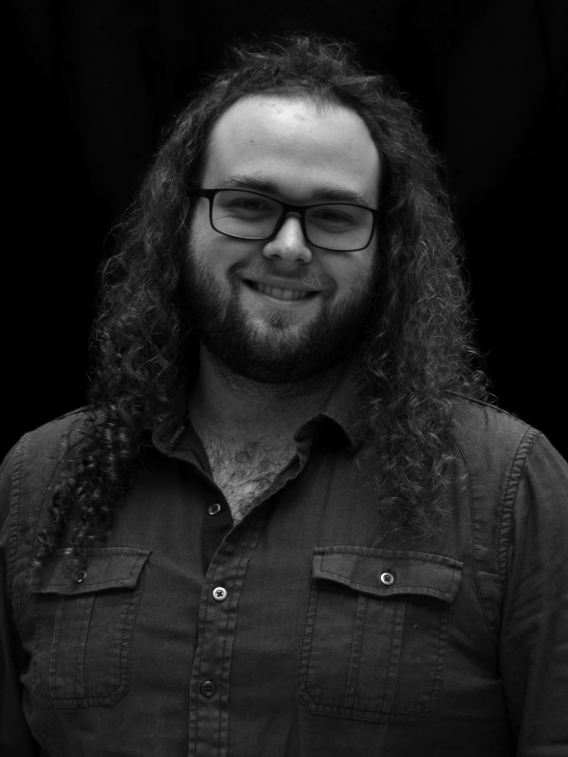 A man with long, curly hair, beard, and glasses smiling at the camera, wearing a button-up shirt, against a dark background.