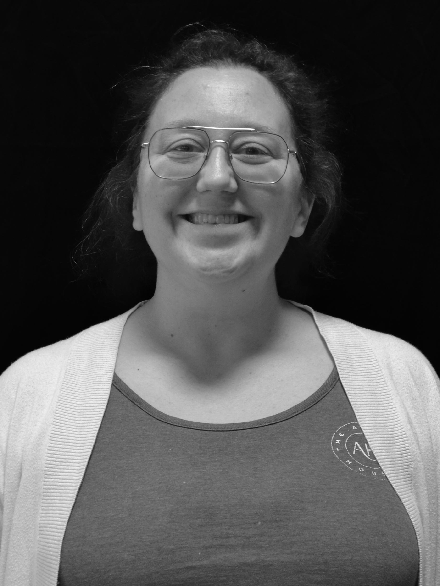 Smiling woman with glasses, wearing a t-shirt and cardigan, against a dark background.