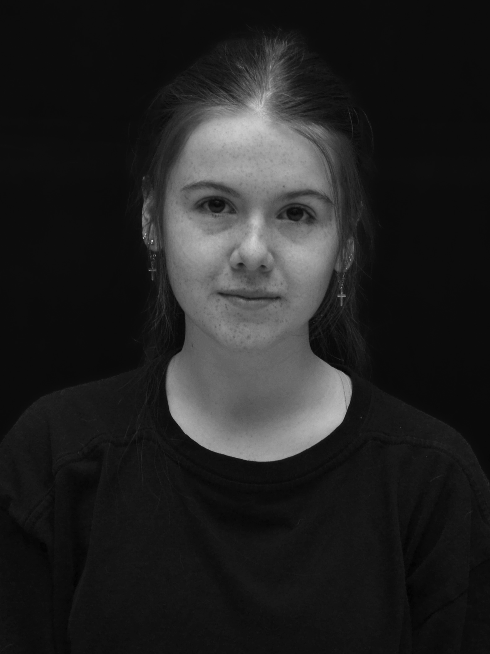 A young girl with freckles and long hair, wearing earrings with a cross design, facing forward against a dark background.