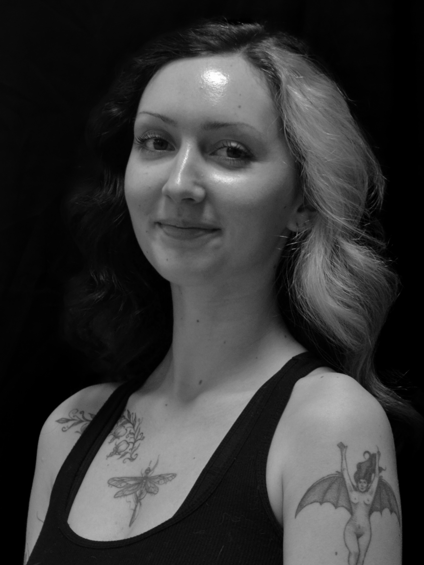 A young woman with wavy hair and tattoos on her shoulders, wearing a black tank top, smiling at the camera.
