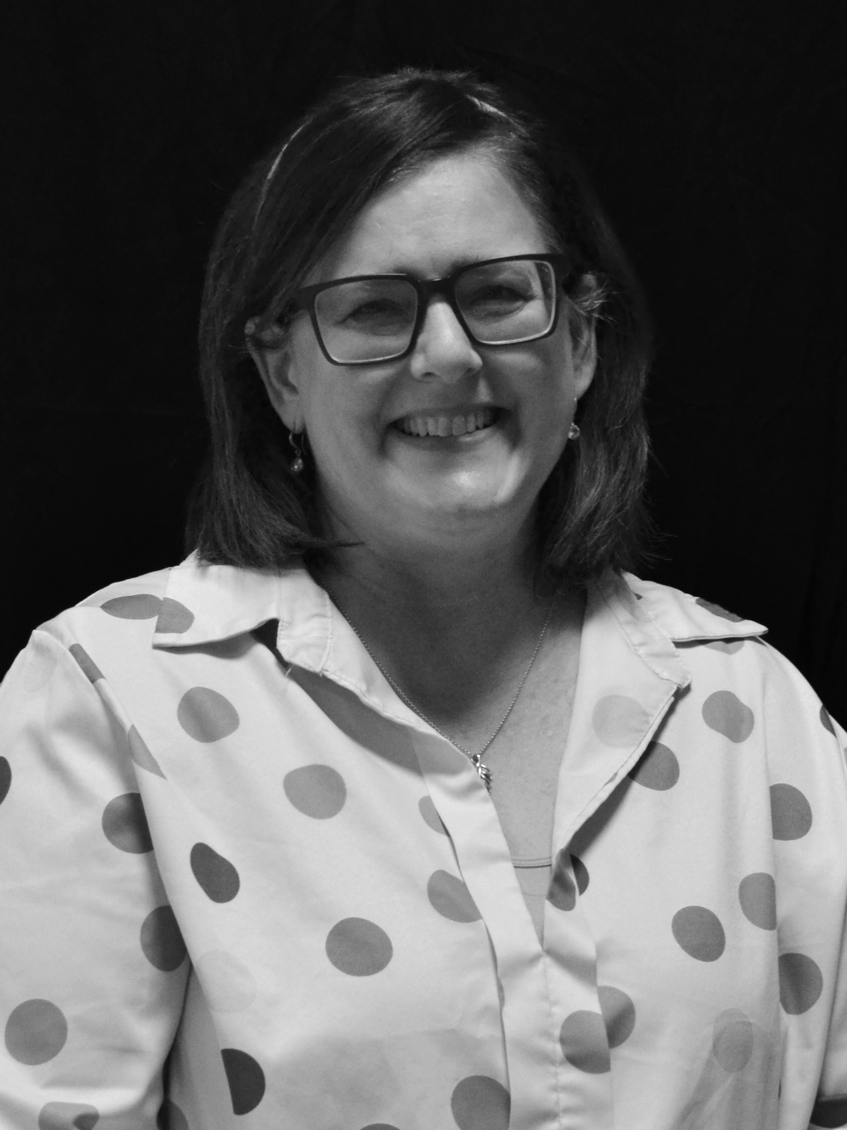A woman smiling, wearing glasses and earrings, with shoulder-length hair, dressed in a polka dot blouse, on a dark background.