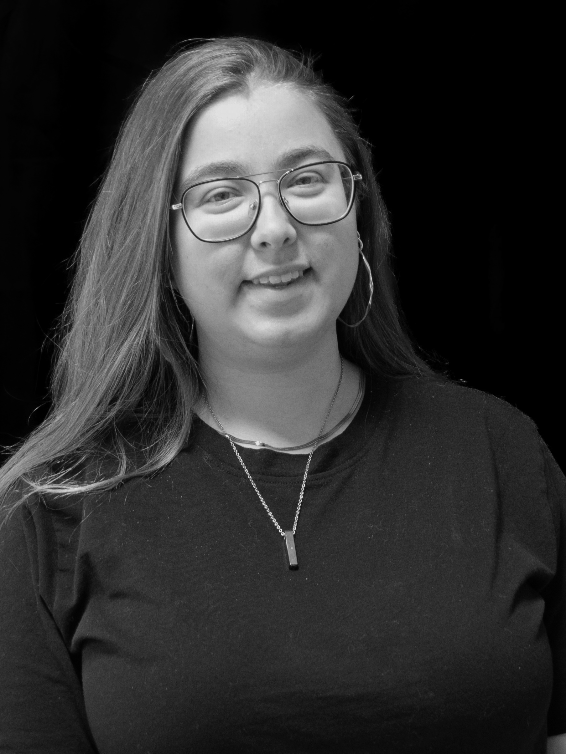 A young woman with long hair wearing glasses, earrings, a necklace, and a black shirt, smiling against a dark background.