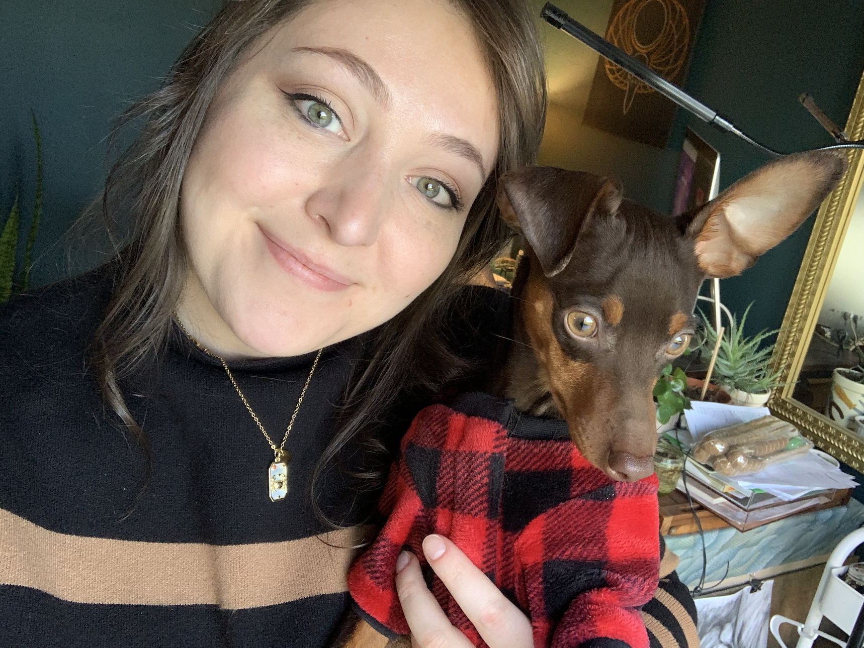A young woman with light skin, long brown hair, and green eyes smiling while taking a selfie with her pet dog. The dog is wearing a red and black checkered sweater and has large ears, and is being held close by the woman in a room decorated with plants, a mirror, and miscellaneous items on a table.