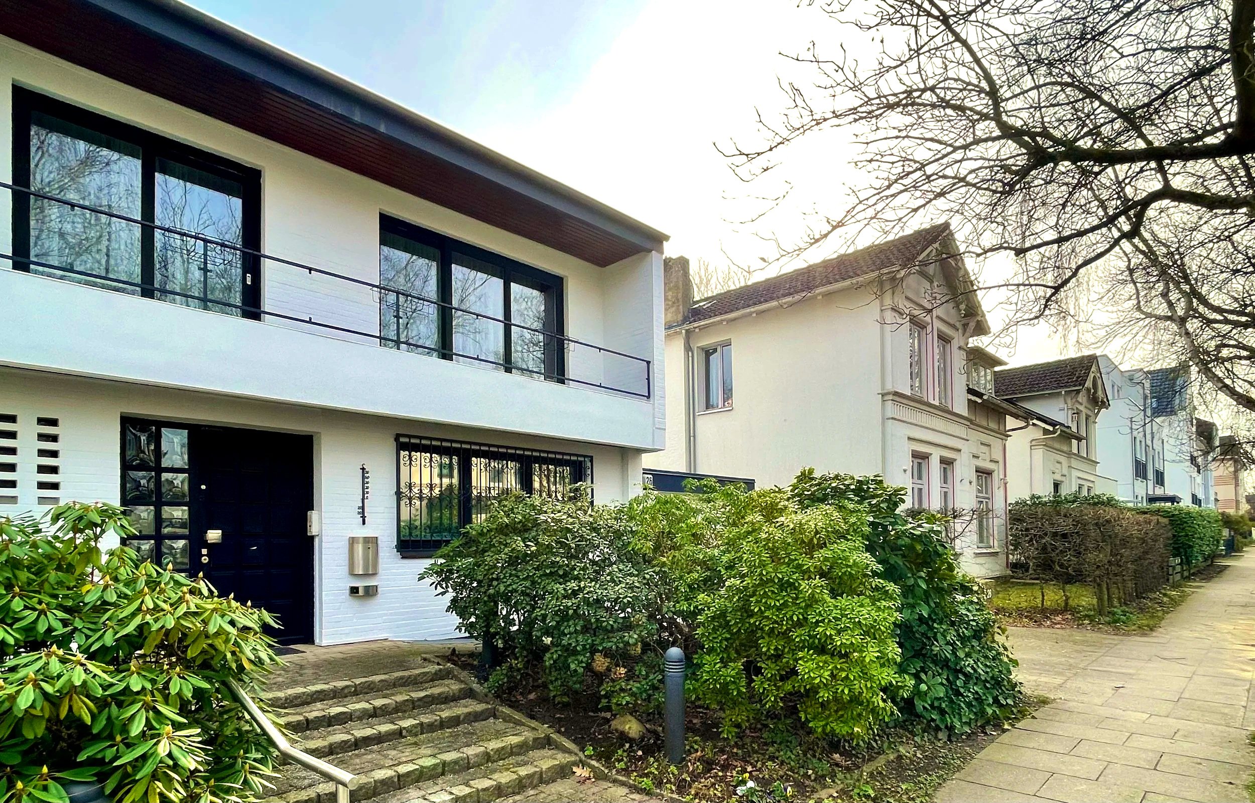 Wohnhäuser in einer ruhigen Straße, modern im Vordergrund und ältere im Hintergrund, mit Büschen und Gehweg, im Sonnenschein.