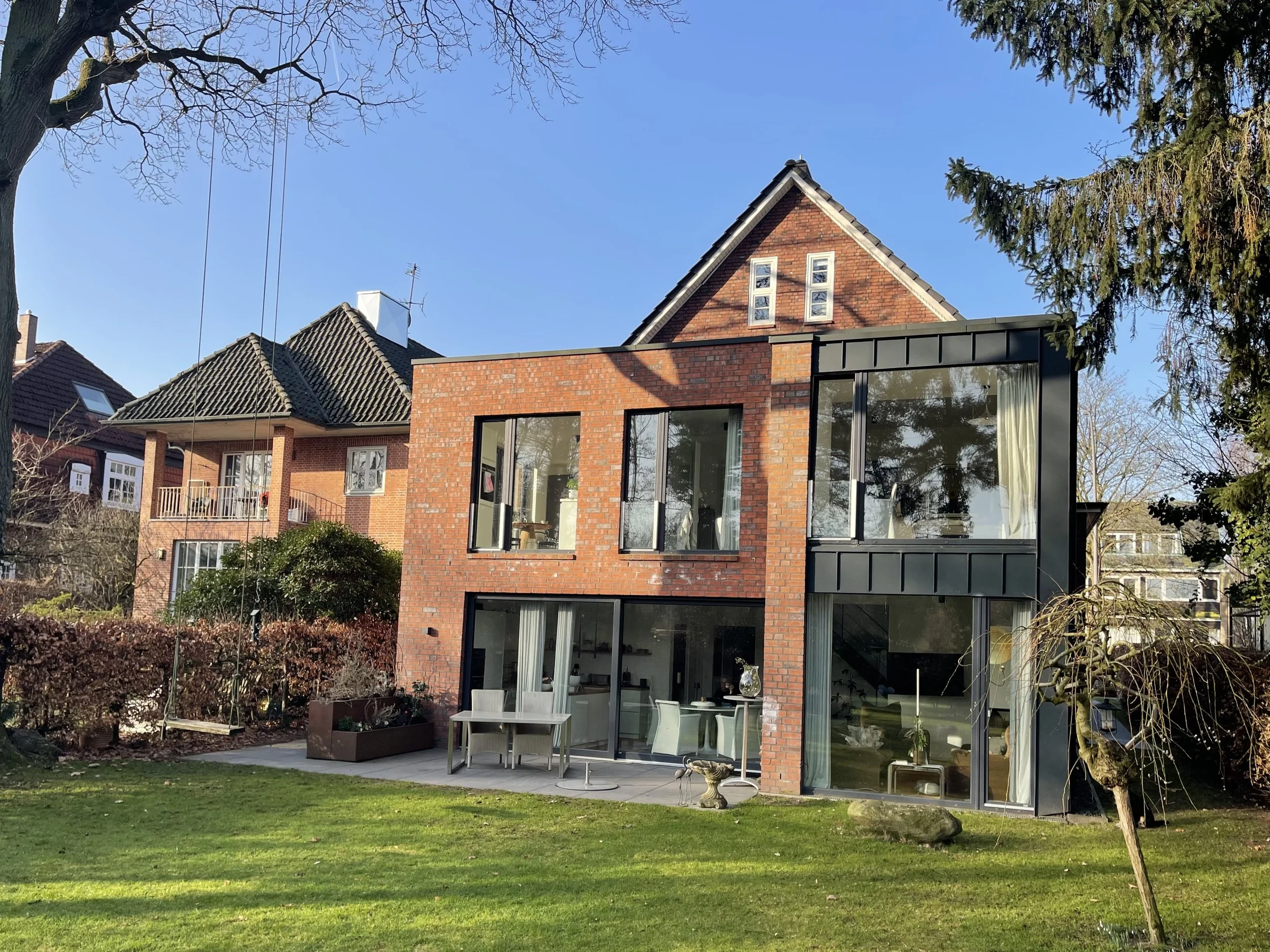 Modernes zweistöckiges Haus mit großen Fenstern und einer Terrasse im Garten. Im Vordergrund ist eine grüne Wiese, links und rechts Bäume, der Himmel ist klar und blau.