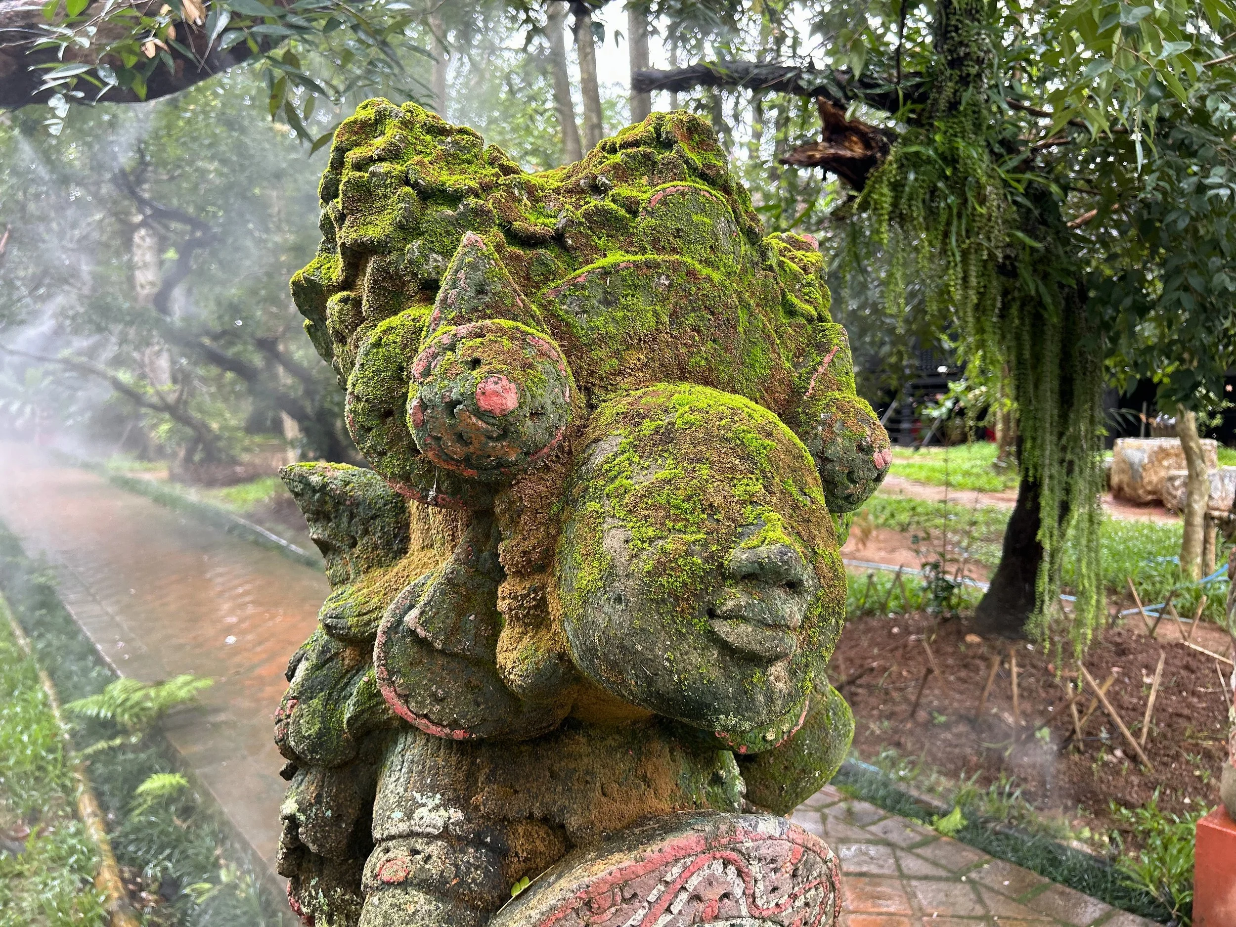 Close-up of a moss-covered stone sculpture of a mythical creature in a lush green garden with trees and plants in the background.