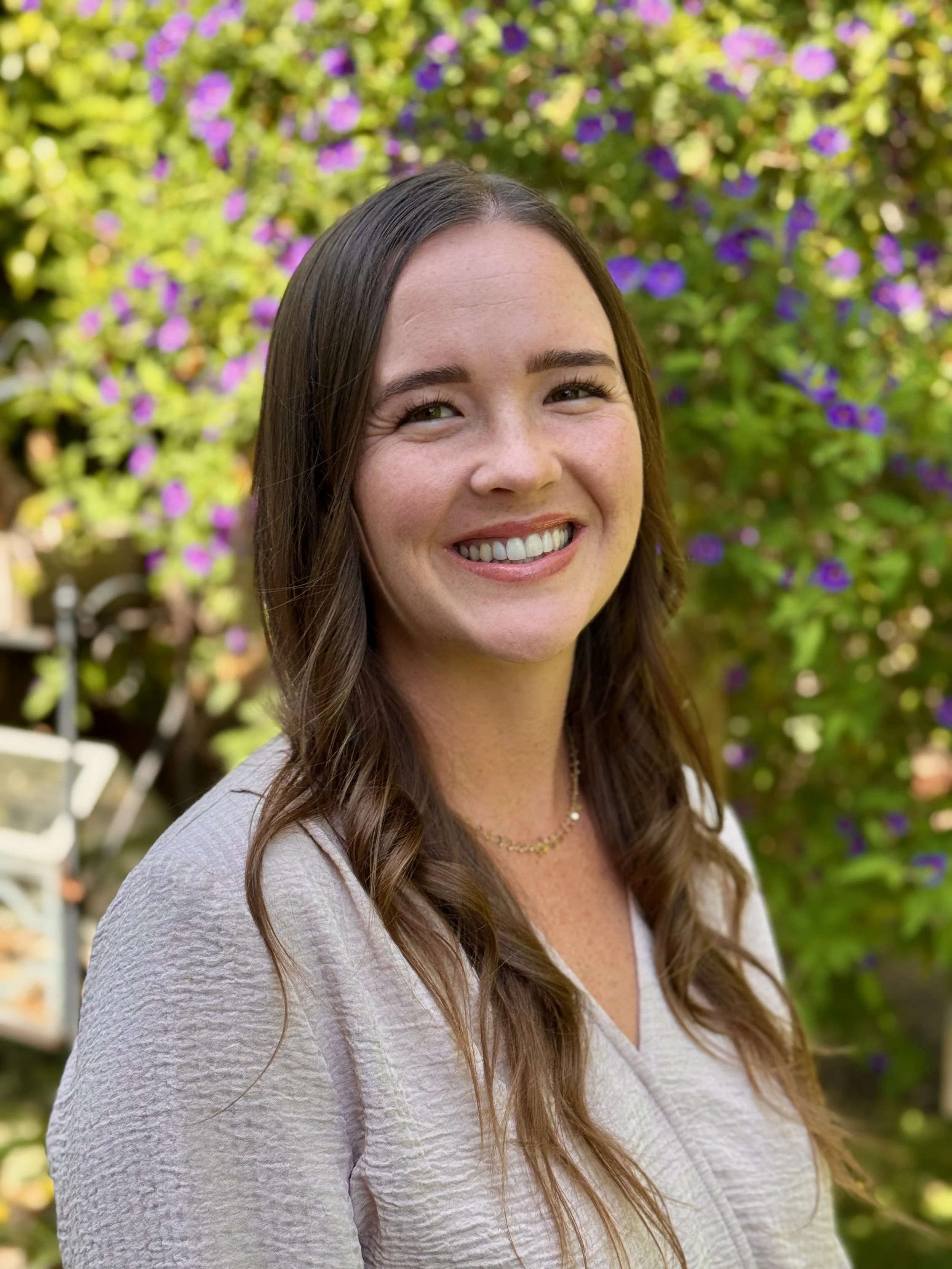 A woman smiling outdoors with purple and yellow flowers in the background.