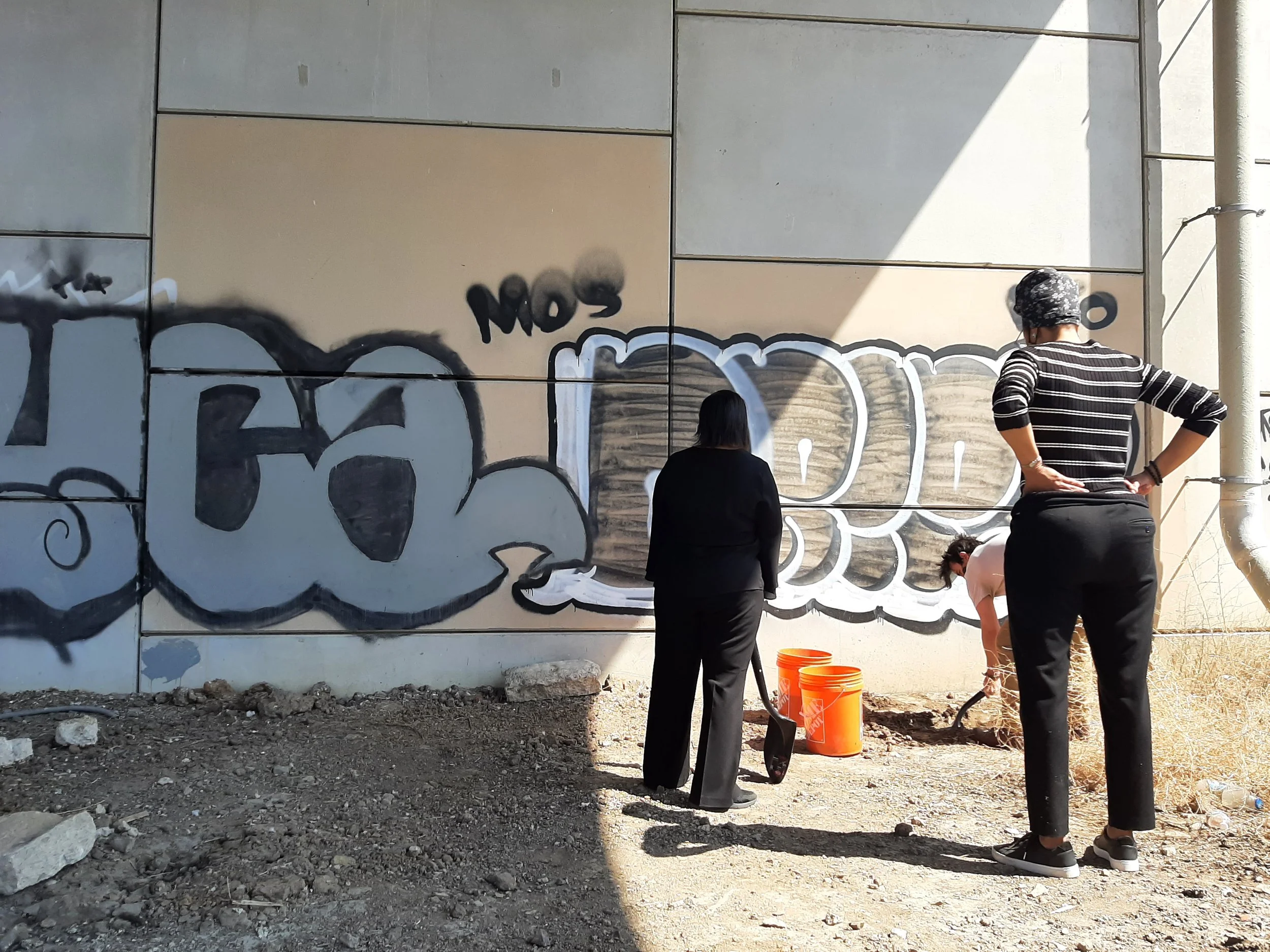 Three women collecting soil from the lynching site of Levi Harrington under a modern bridge with grafitti in the background.