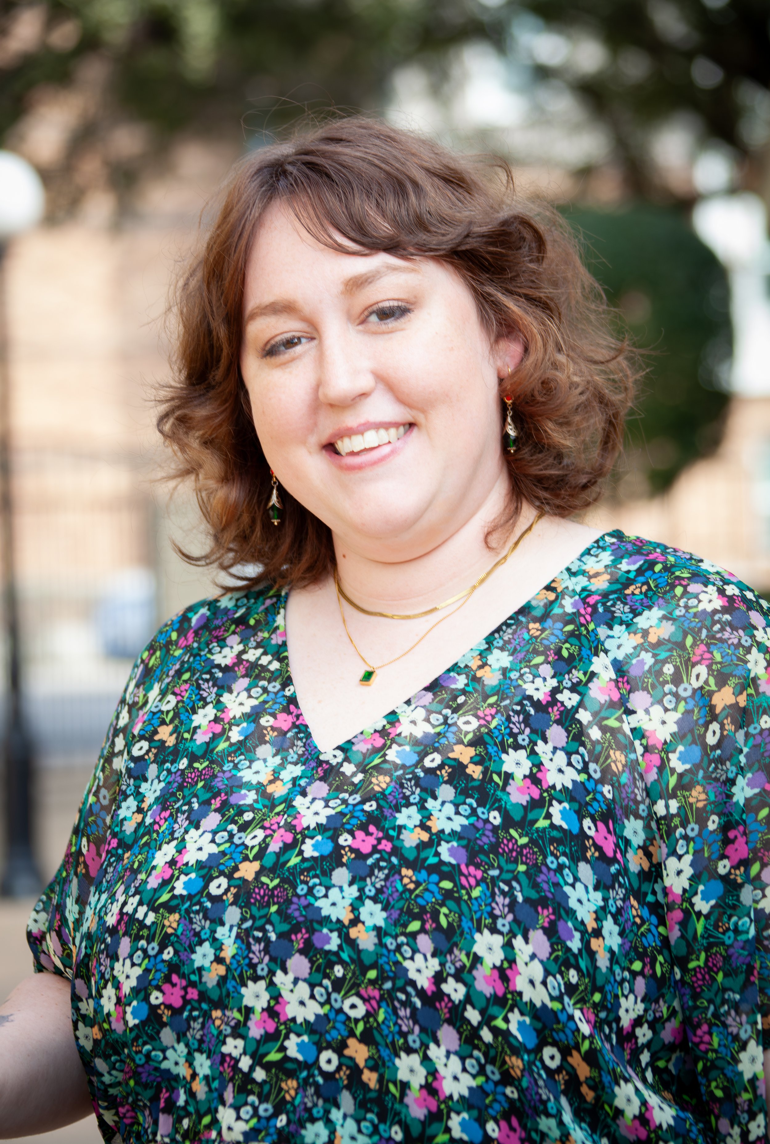 Portrait of Aria, smiling at the camera in a floral dress and gold jewelry.