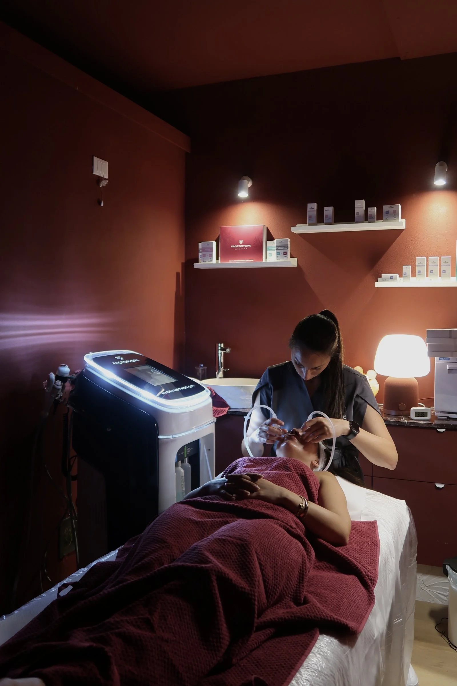 Una donna che riceve un trattamento di estetica o di terapia sul volto di un cliente, sdraiato su un lettino, in una stanza con pareti di colore rosso scuro e scaffali con prodotti di bellezza. Sulla superficie vicino ci sono apparecchiature medicali e una lampada da tavolo accesa.