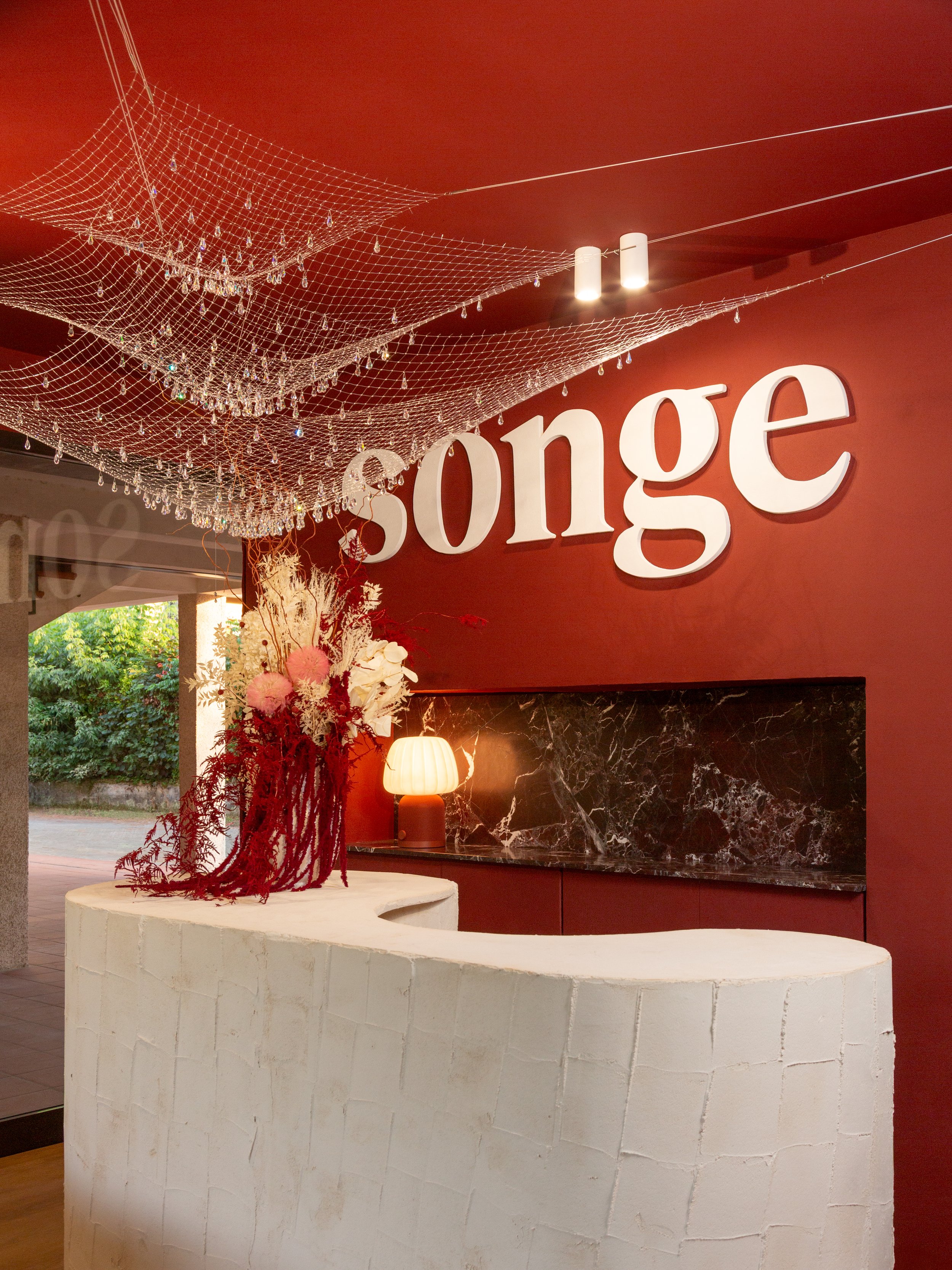 Un rivestimento con la parola 'Songe' in lettere bianche 3D su una parete di colore rosso scuro; nel primo piano, un'installazione floreale con fiori in tonalità rosa, bianco e rosso, e una lampada di forma rotonda sopra un bancone bianco con una superficie di pietra scura.