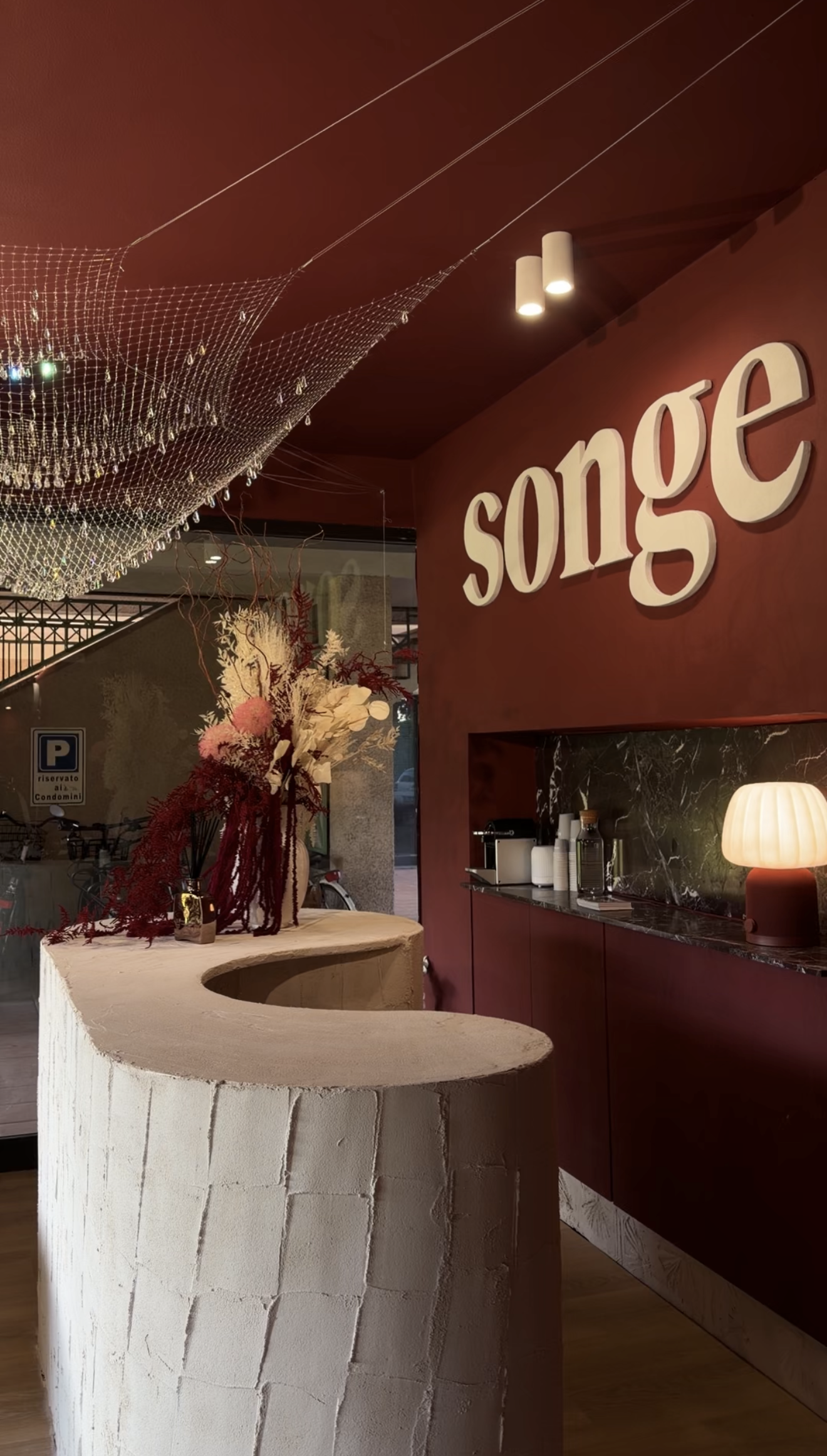 Interno di un locale con parete rossa, decorato con una grande scritta 'song' in rilievo. Vi è un bancone con finitura in marmo nero e vari oggetti, tra cui bottiglie e una lampada con paralume rotondo e caldo.
