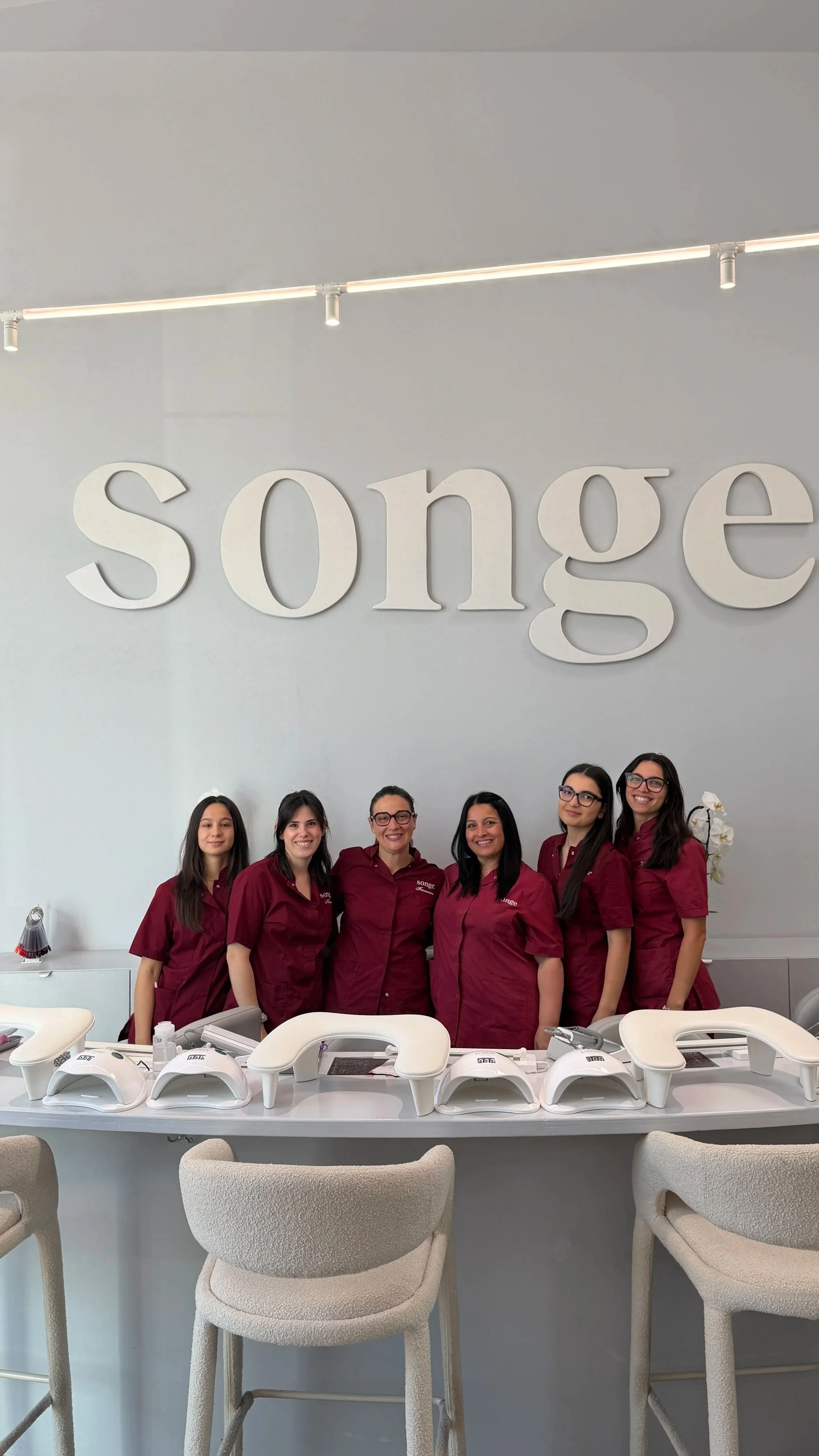 Cinque donne sorridenti indossando divise rosse e che stanno in un ufficio di bellezza, con strumenti per manicure sulla scrivania.